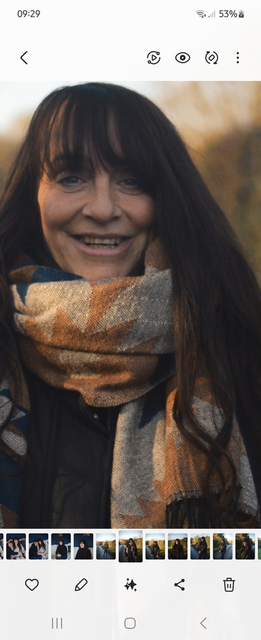 A woman with long dark hair smiling outdoors during golden hour, wearing a patterned scarf and a dark jacket.