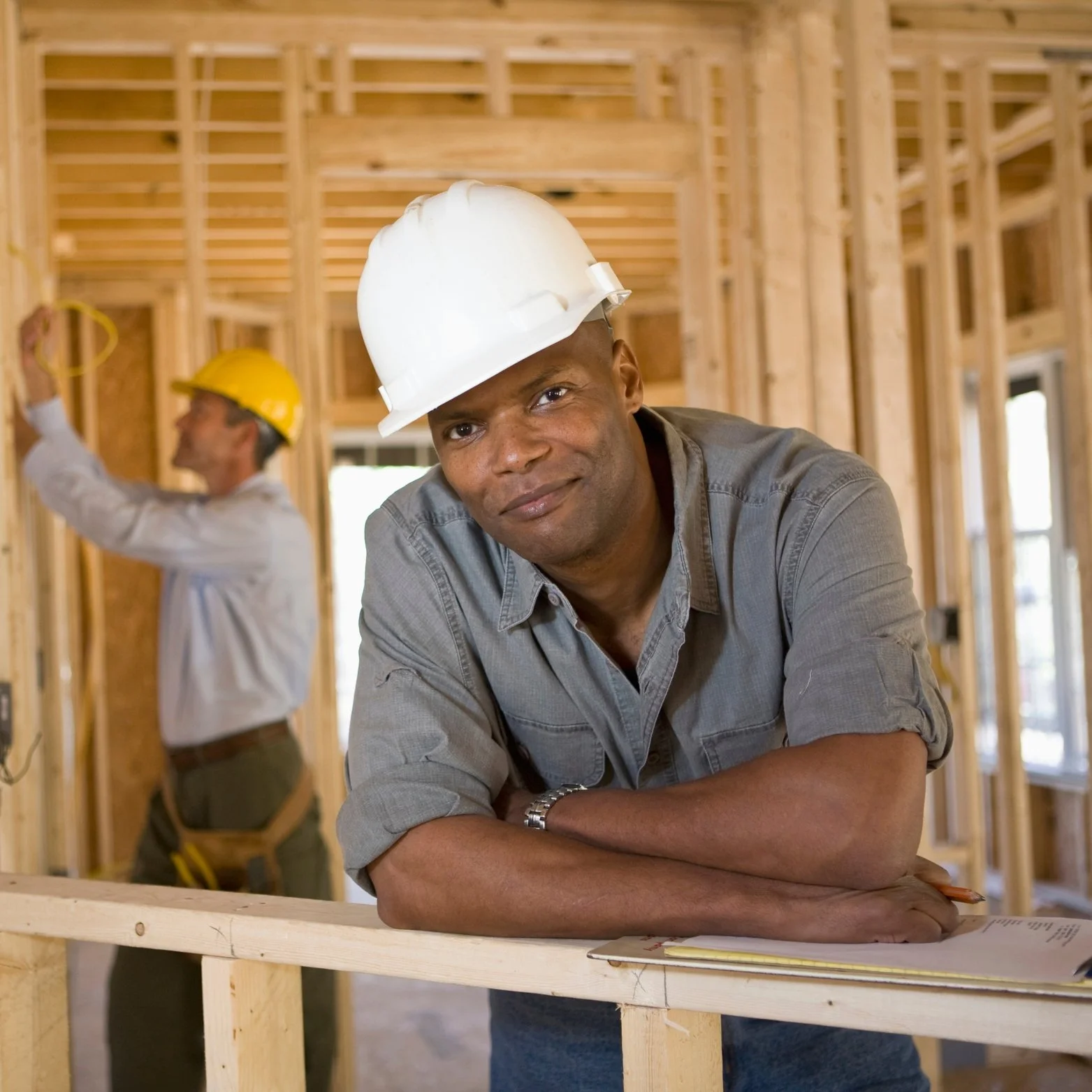 A man wearing a white hard hat leaning on wooden framing in a house under construction, with another worker in a yellow hard hat working in the background.