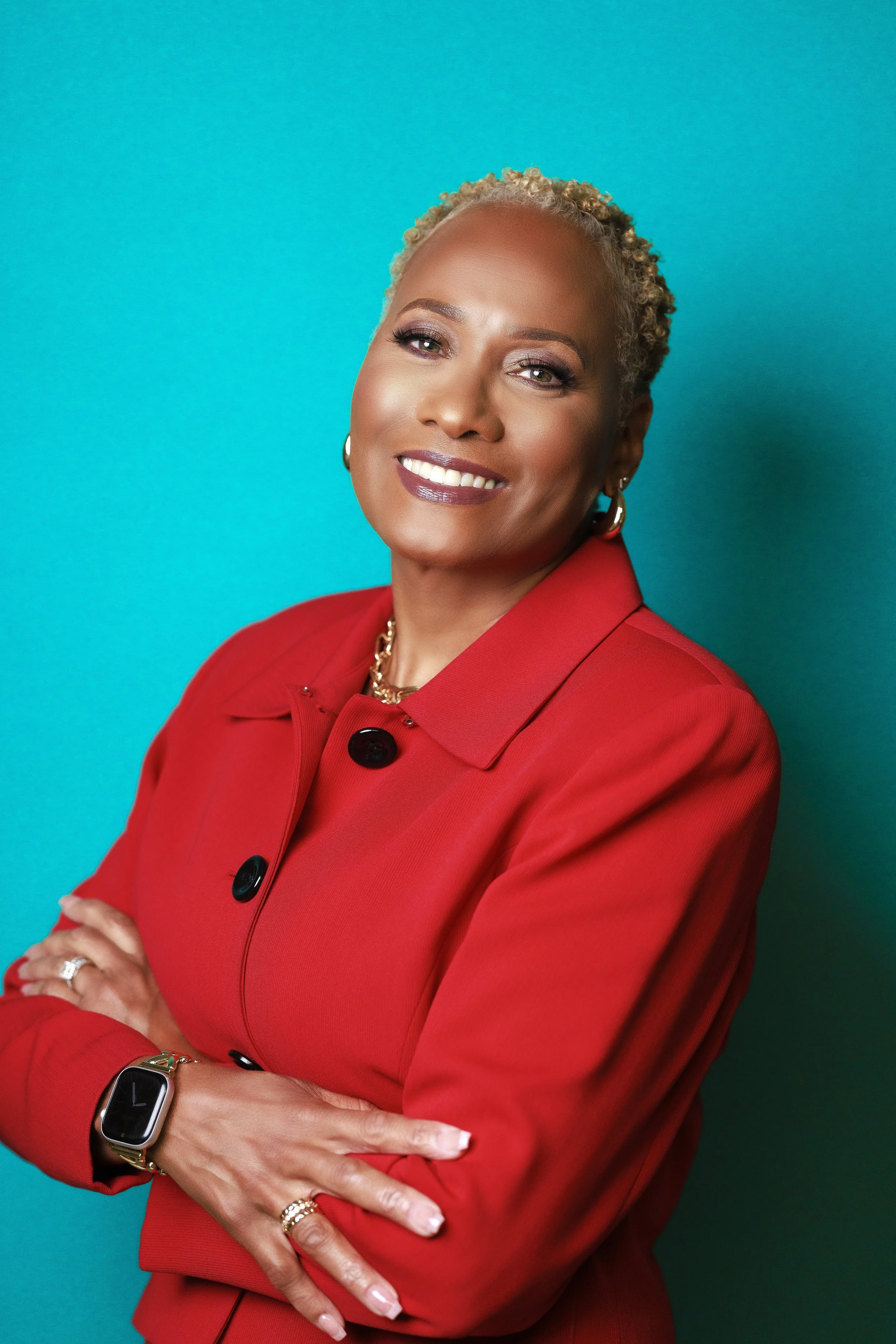 A confident woman with short curly blonde hair, wearing a red blazer, standing with arms crossed against a teal background, smiling.