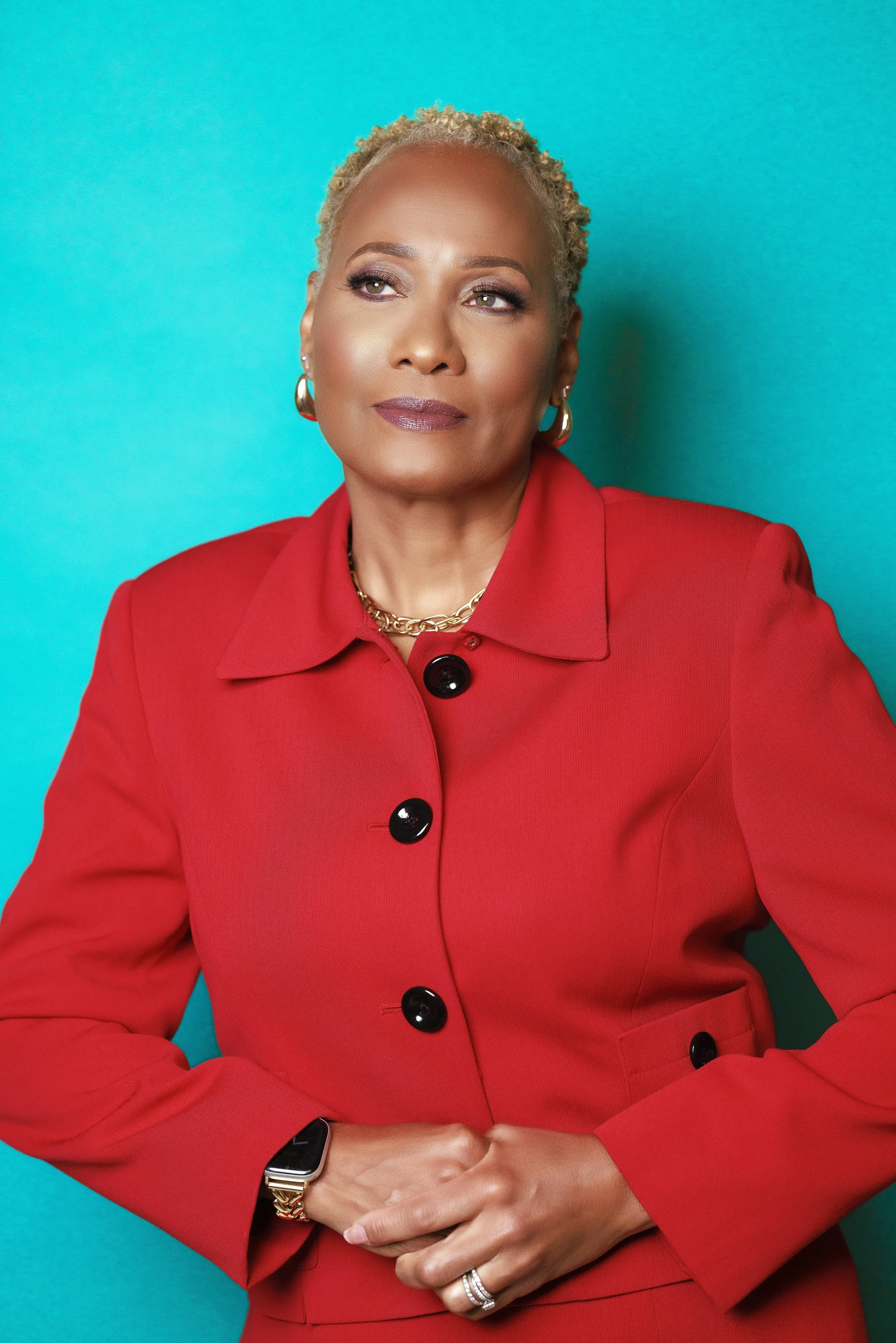 Portrait of a confident woman with short blonde hair, wearing a red blazer and jewelry, against a vibrant blue background.