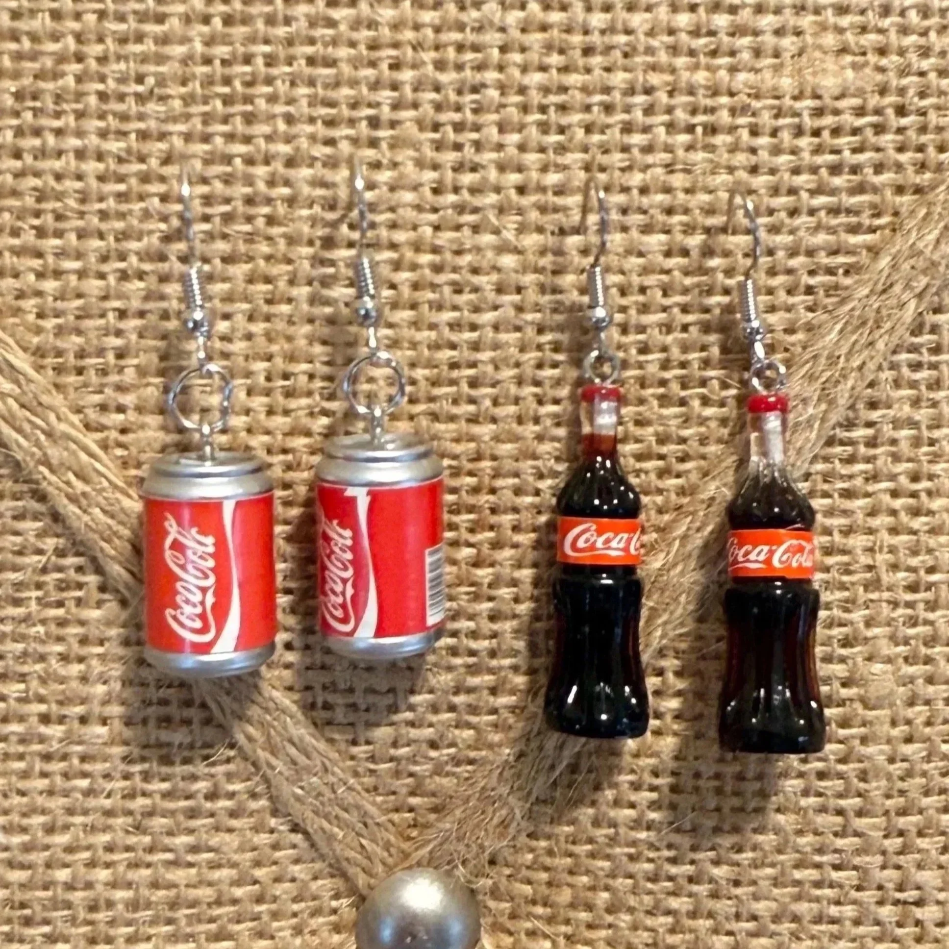 Earrings shaped like miniature Coca-Cola cans and bottles hanging on a burlap background.