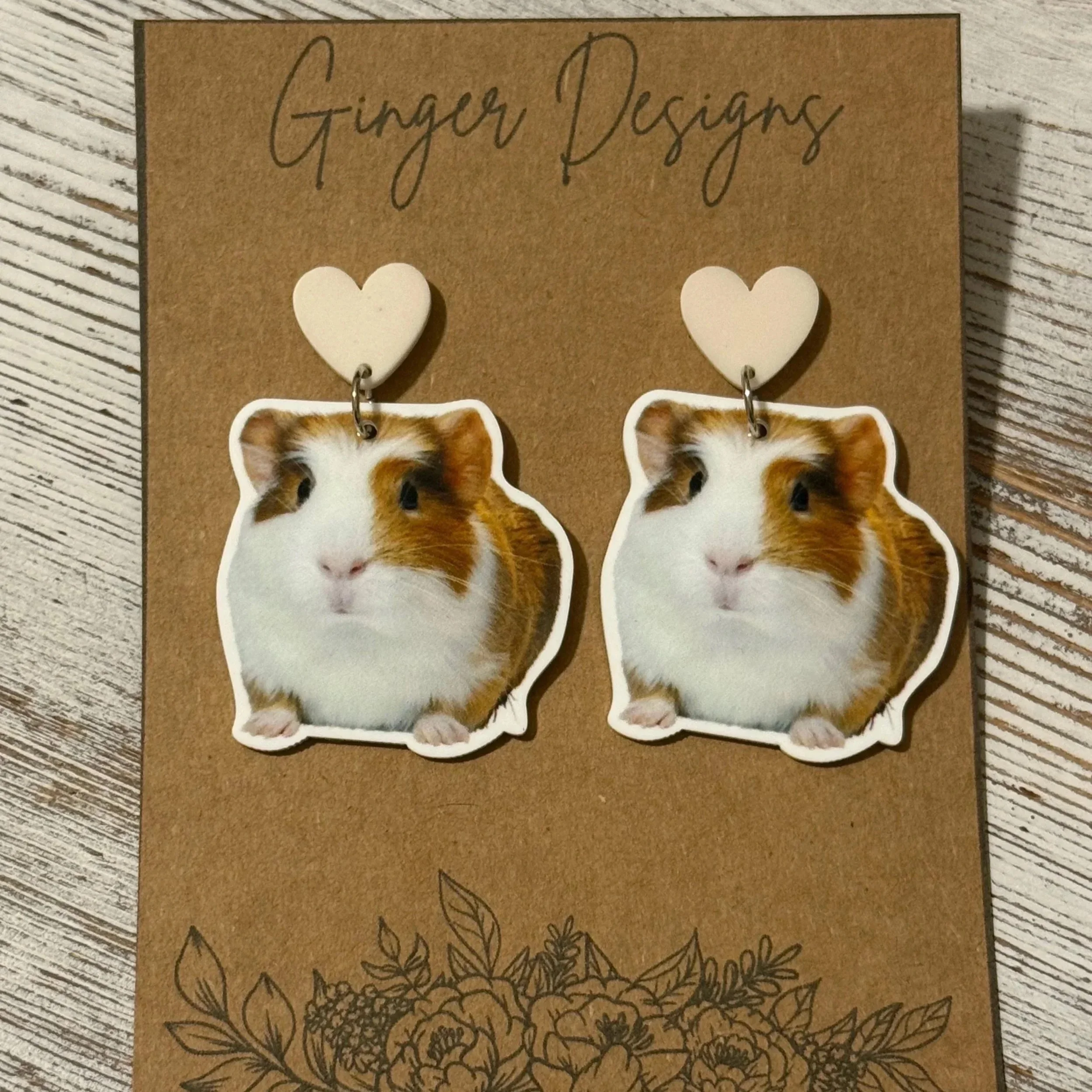 Pair of guinea pig-shaped earrings with white heart connectors on a brown card. The earrings feature a guinea pig with white, brown, and black fur, and are displayed on a wooden surface.