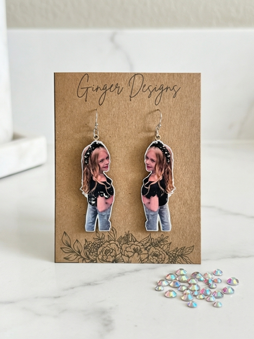 Custom earrings featuring a young girl's photo on a brown card labeled 'Ginger Designs,' with a floral illustration at the bottom, and small iridescent rhinestones nearby.