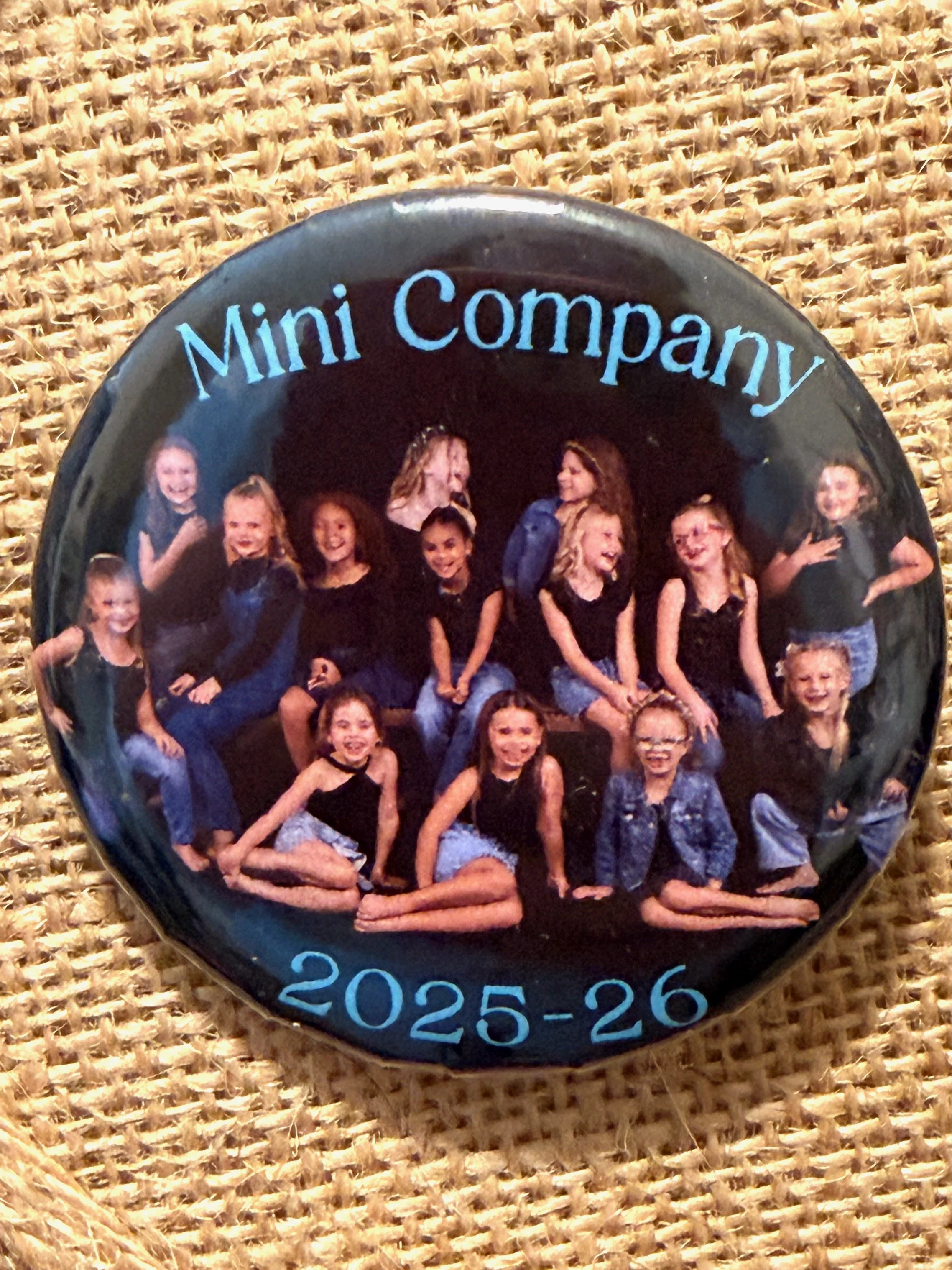 A round button featuring a group of girls posing together, with the words 'Mini Company' at the top and the years '2025-26' at the bottom. The girls are sitting and standing, smiling and dressed casually.