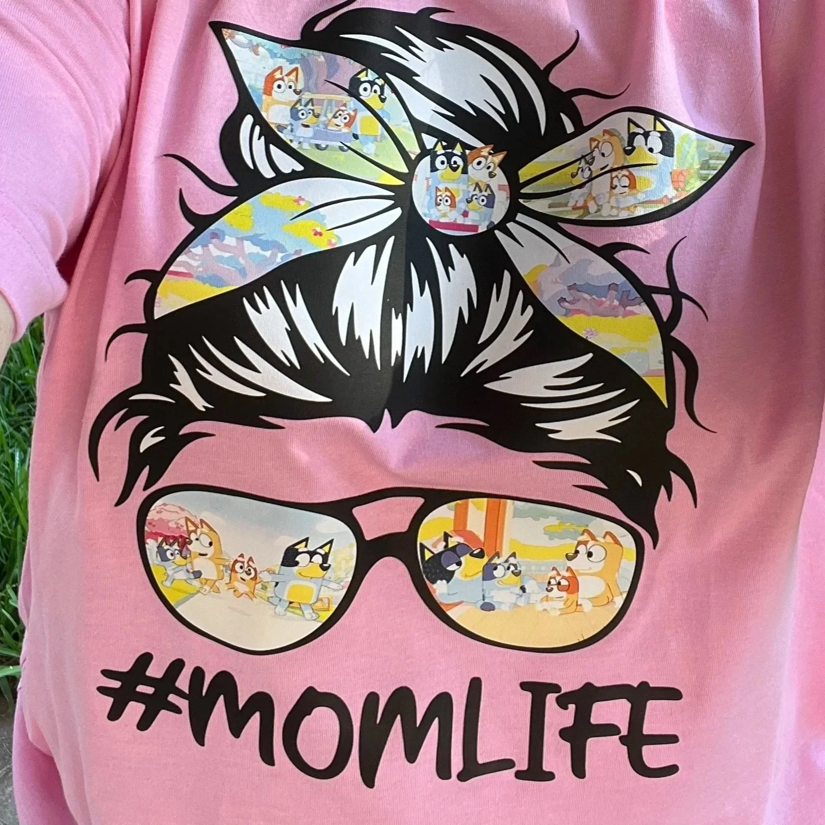Pink T-shirt with a graphic of a mom’s head with hair tied back and large glasses showing scenes from the cartoon “Bluey” inside the hair and glasses, and the hashtag #MOMLIFE.