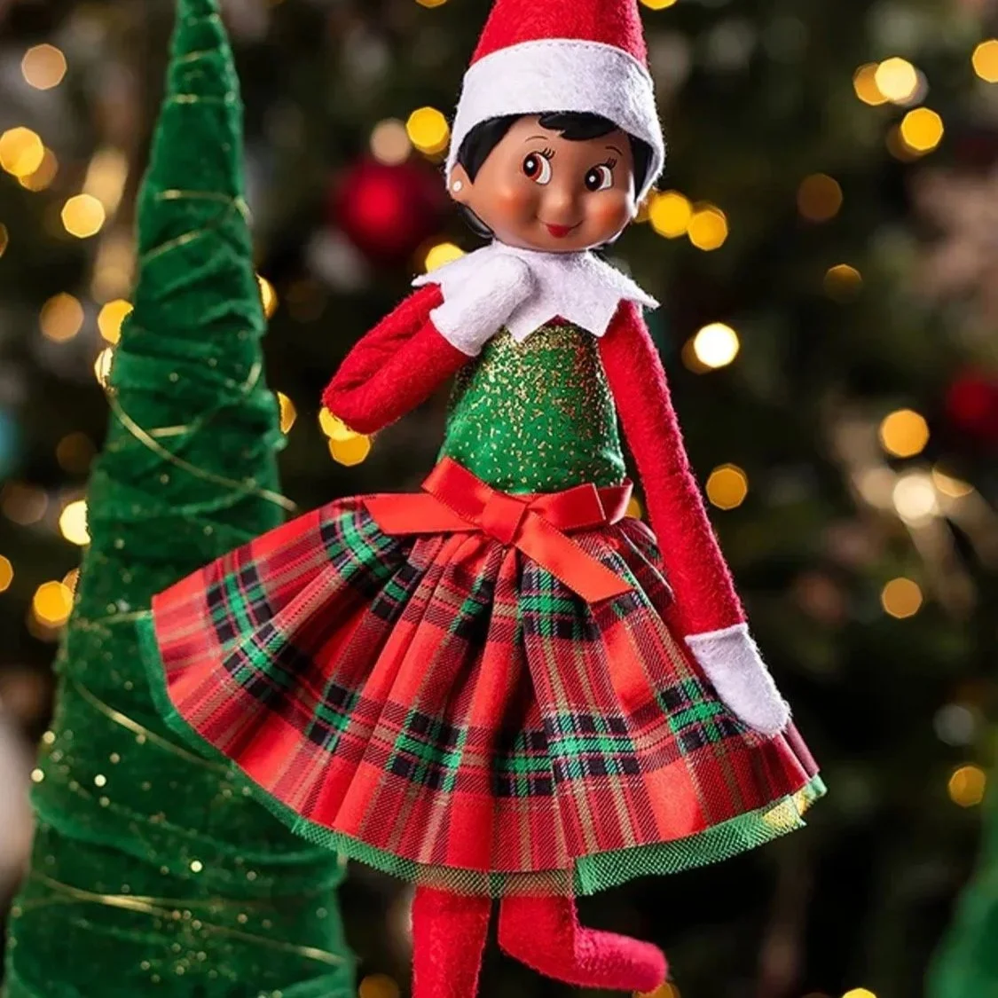 Christmas elf doll wearing a red and green plaid skirt, a green top with gold glitter, a red Santa hat, and red tights, standing near a green spiral tree decoration on a Christmas tree background.