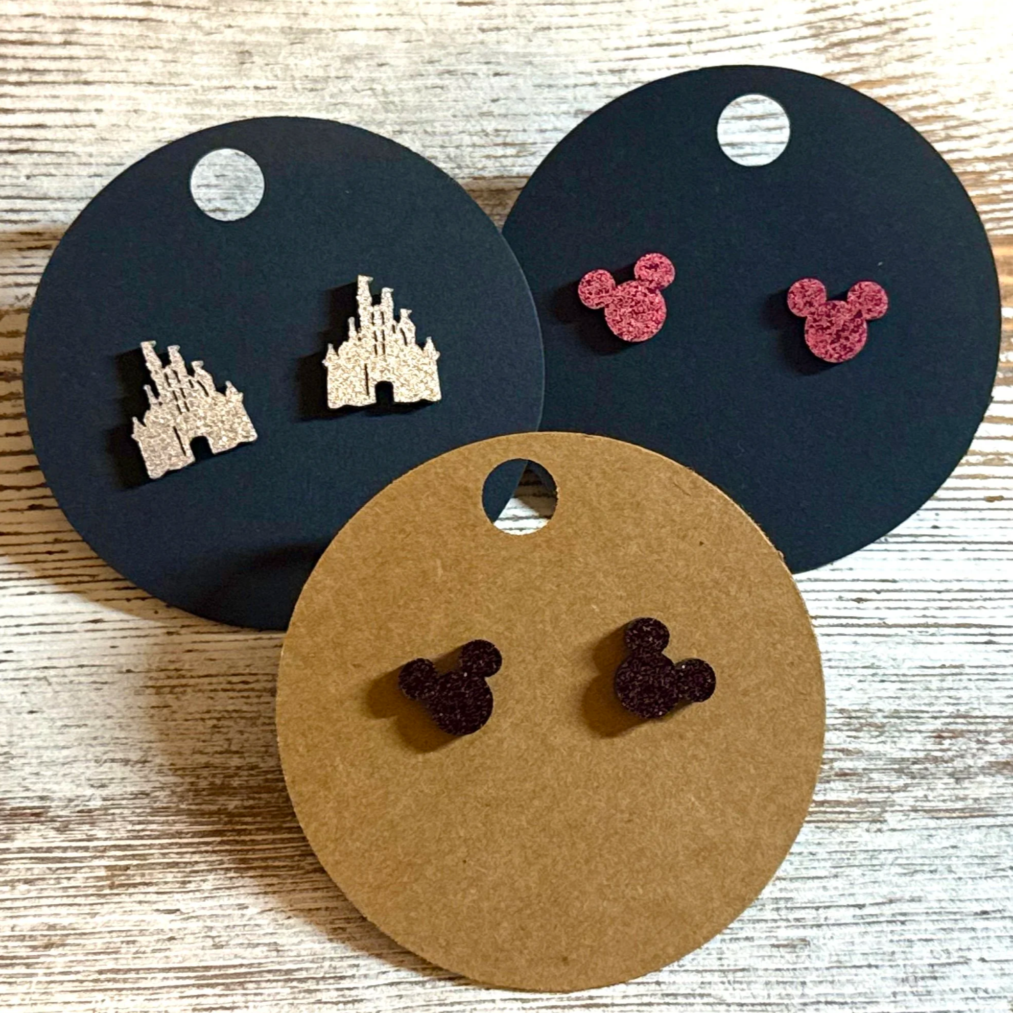 Three pairs of Mickey Mouse-shaped earrings on display, with two pairs on black backing cards and one pair on a brown backing card, featuring glittery pink, glittery dark purple, and glittery silver designs.