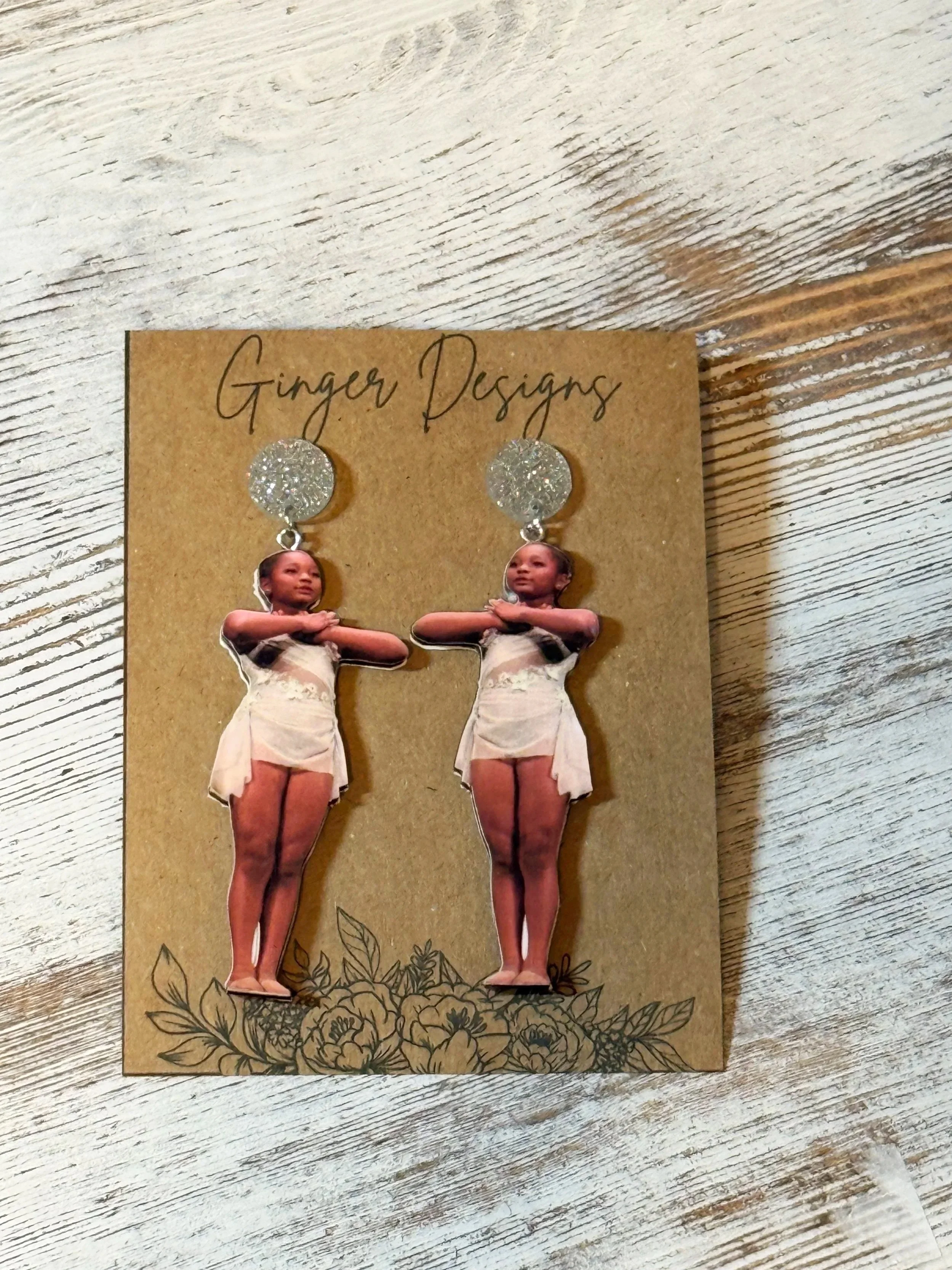 A pair of earrings featuring a cut-out photo of a young girl with arms crossed, standing with legs together, on a brown card with floral illustrations at the top and bottom. The card has the brand name "Ginger Designs" handwritten at the top.