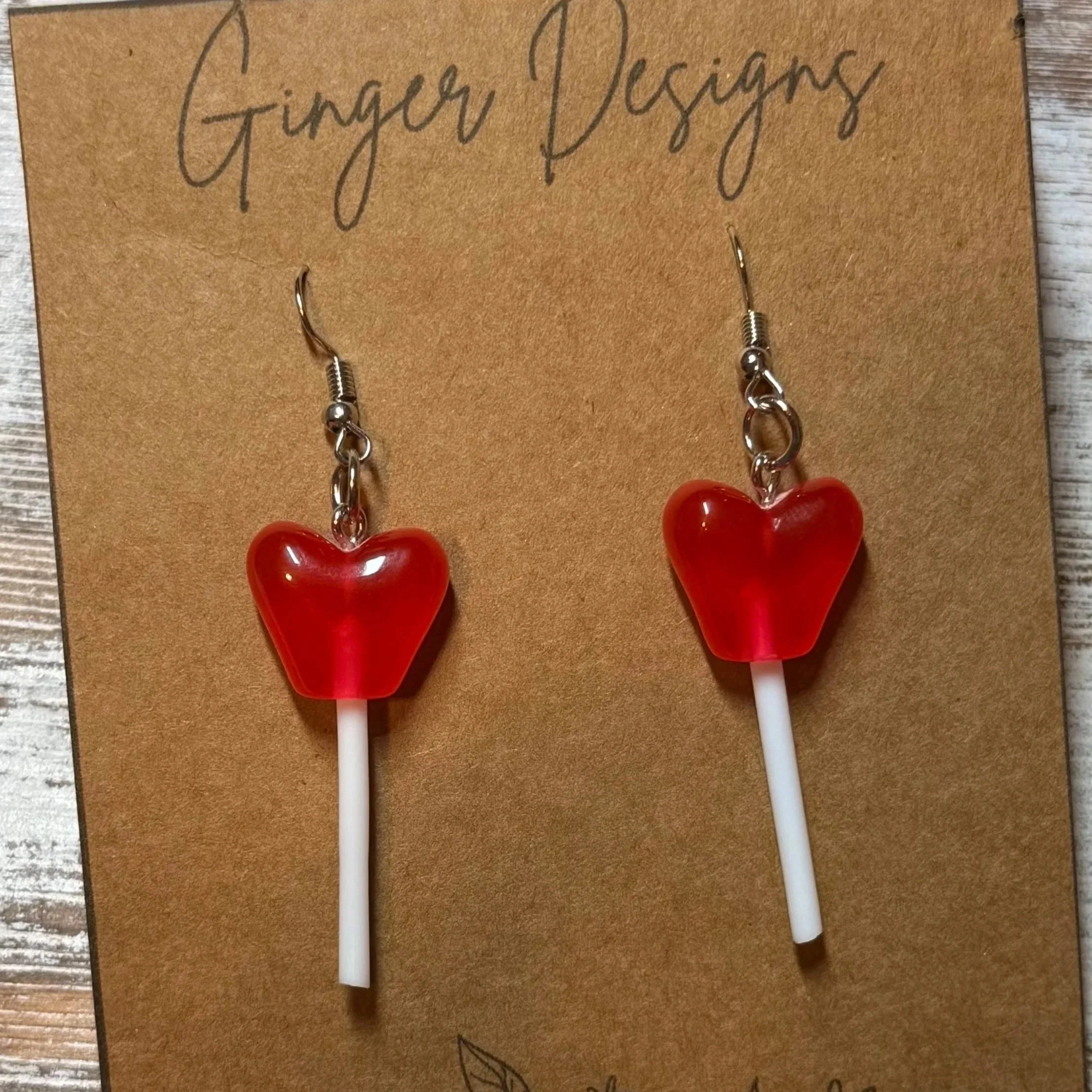 Pair of heart-shaped red lollipop earrings on white sticks with a  tan card labeled 'Ginger Designs'.