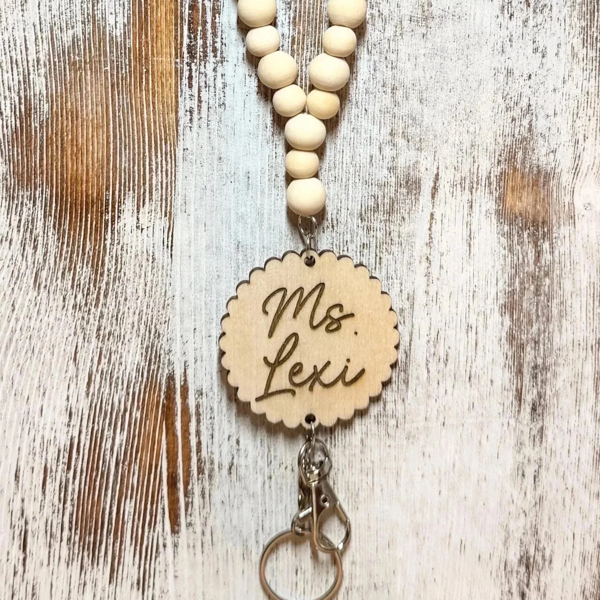 A beaded teacher lanyard with wooden beads and a round wooden tag engraved with the teachers last name hanging on a rustic white wooden surface.