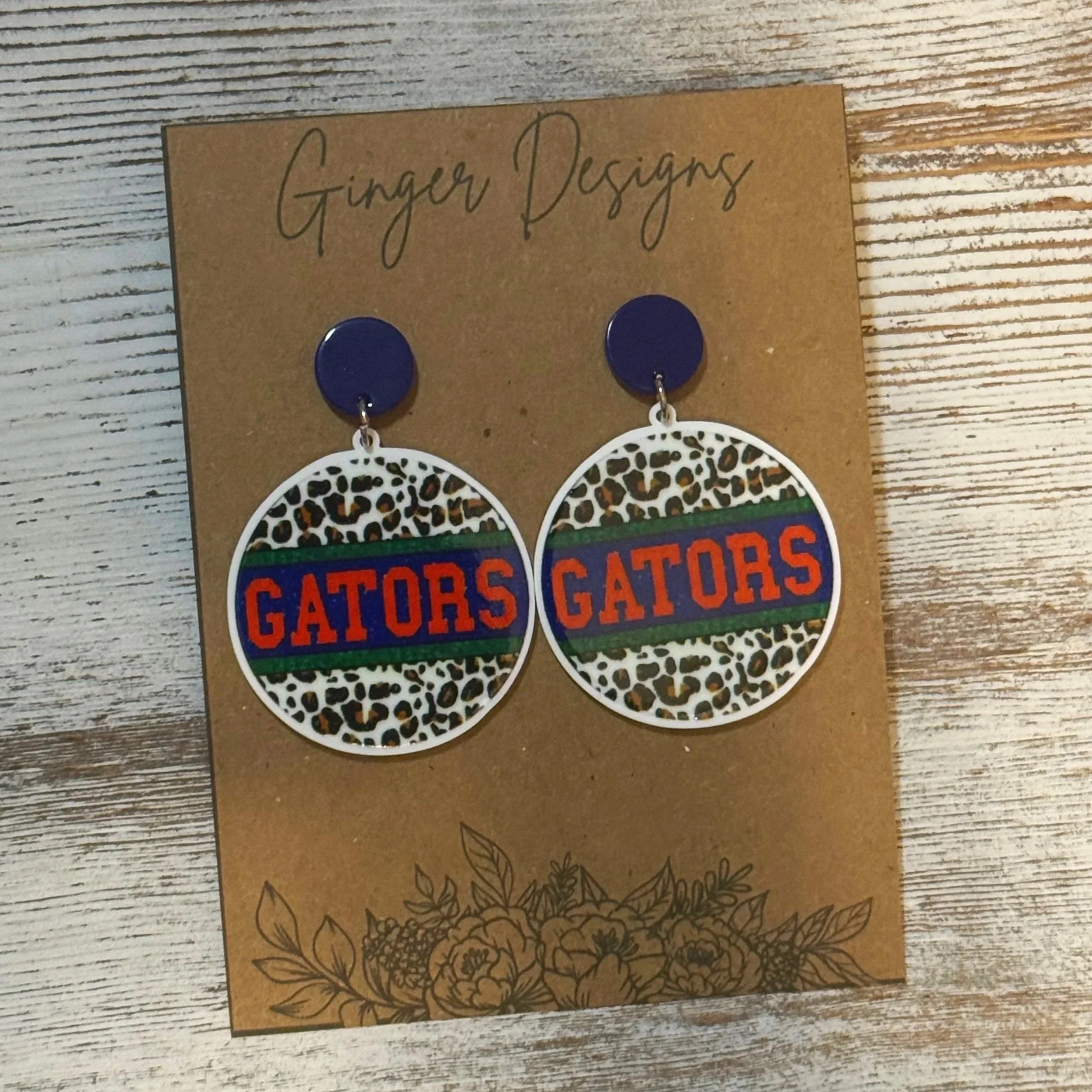 Earrings with leopard print, Gators logo, and blue studs on brown card from Ginger Designs, placed on a wooden surface. Gator Fans.