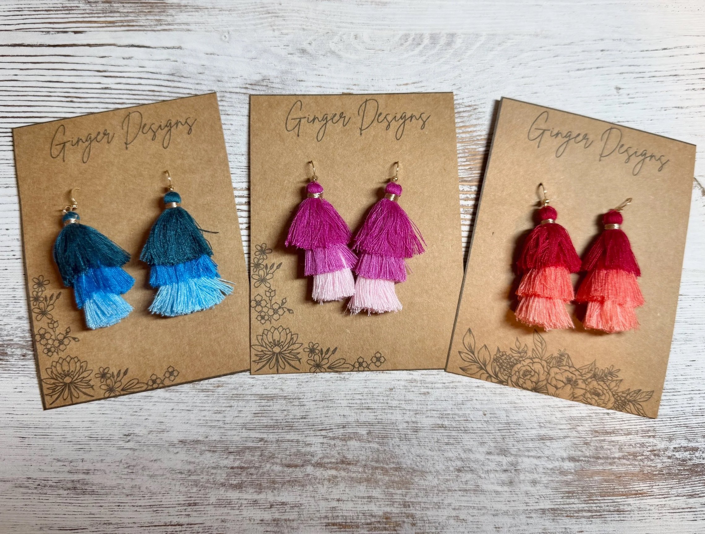 Three pairs of handmade tassel earrings in different colors displayed on kraft paper cards with floral designs, on a white wooden surface.