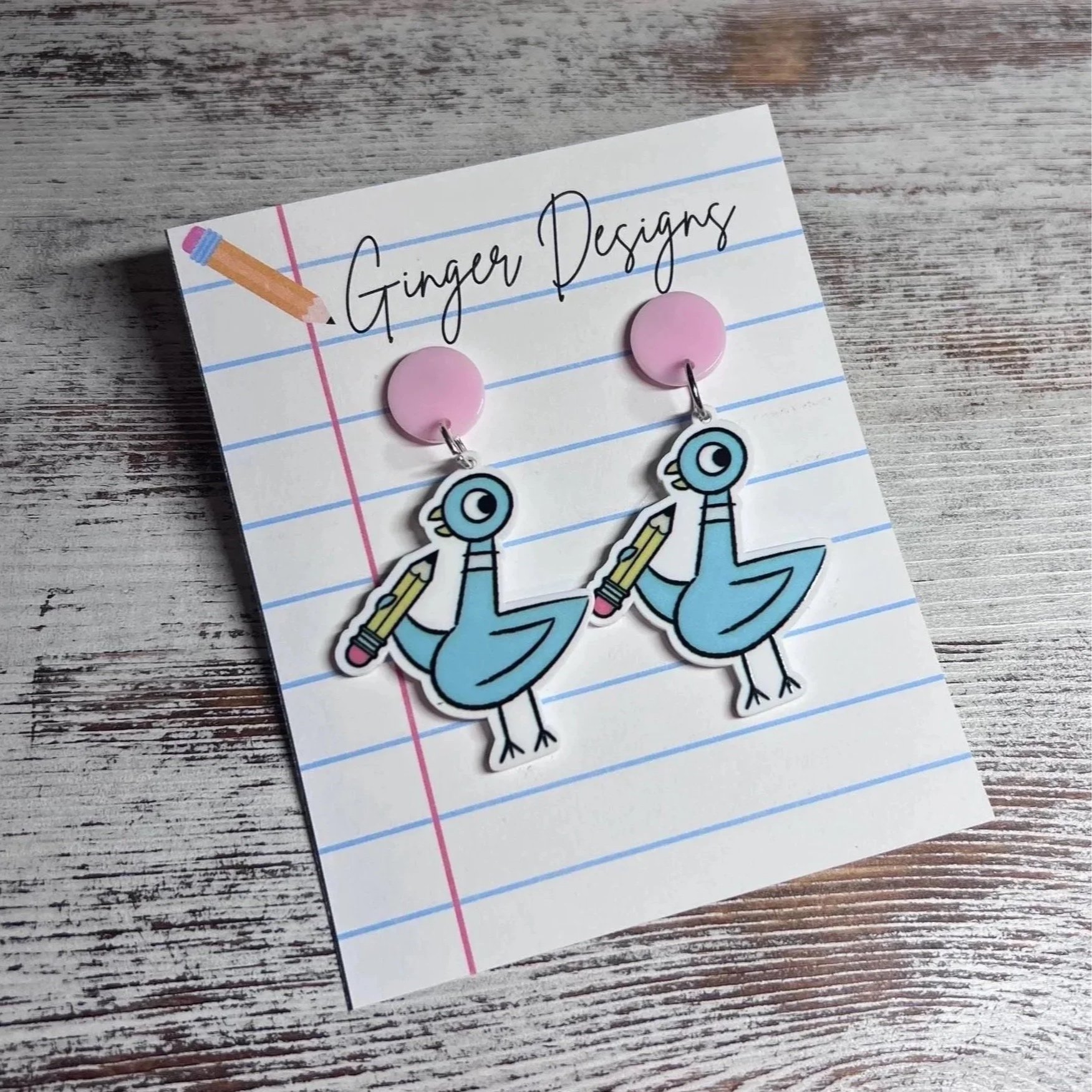 Pair of earrings featuring cartoon character “Pigeon” holding pencils, attached to pink round bases, on a notepad with 'Ginger Designs' written at the top.