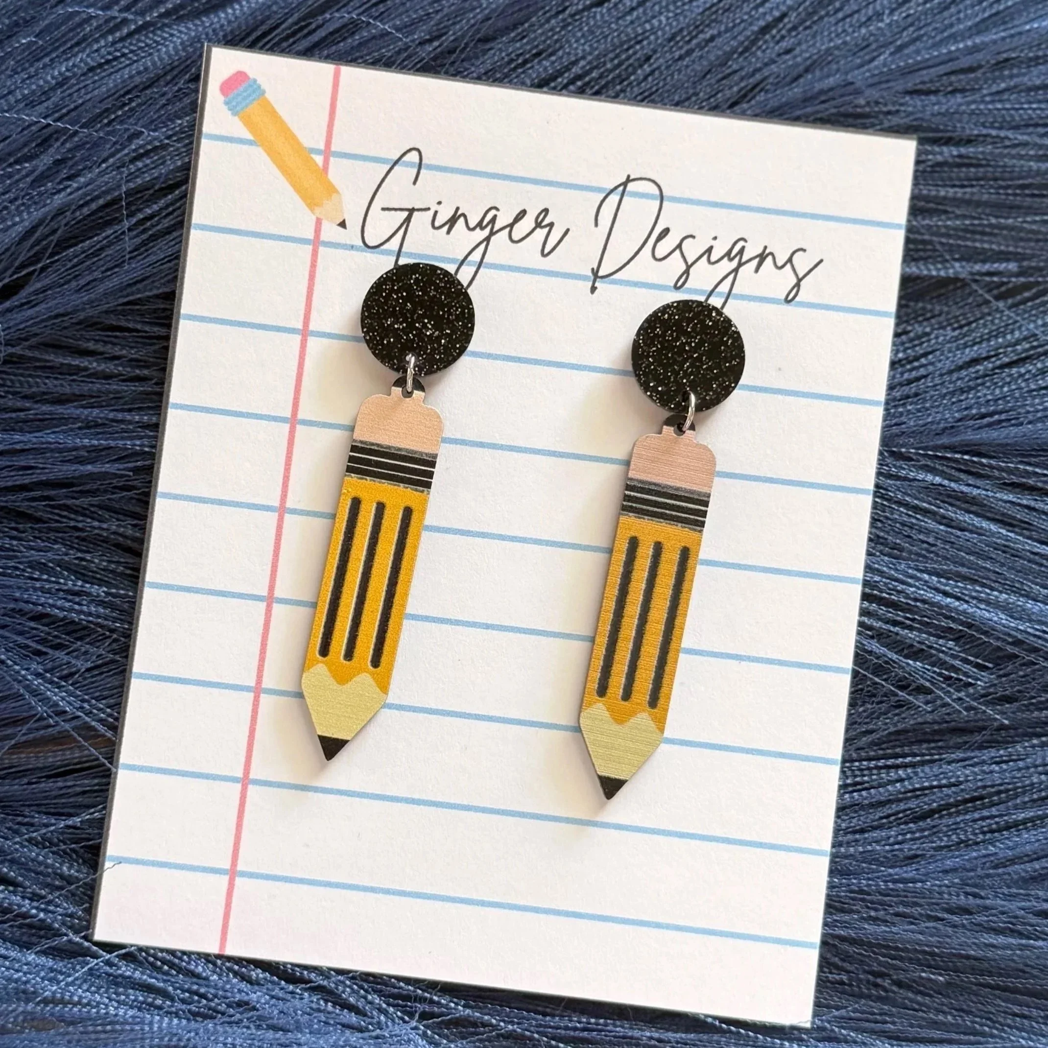 Pair of earrings shaped like pencils on a notecard with the words 'Ginger Designs' written on it, placed on a textured dark blue surface.