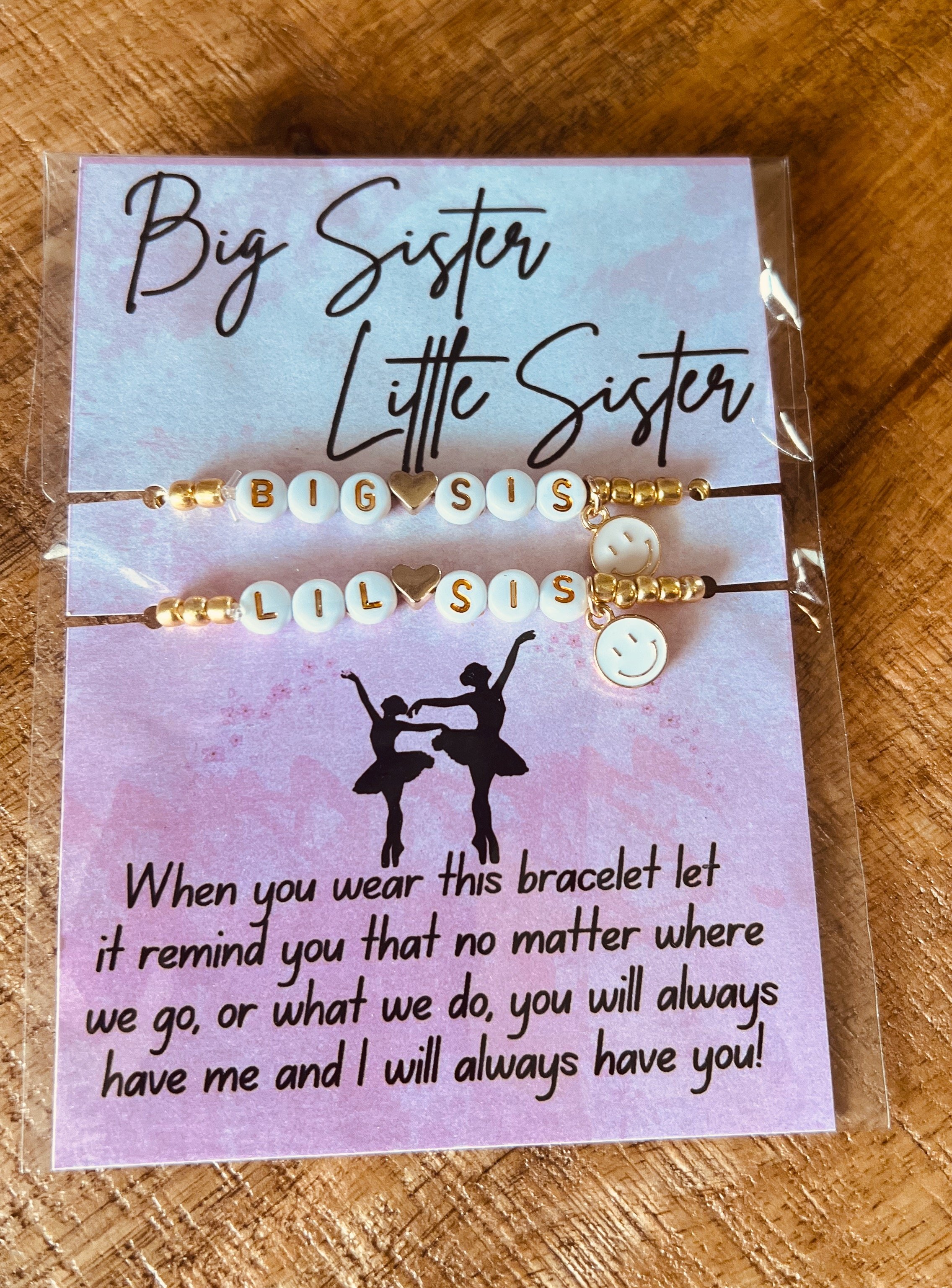 A bracelet with beads spelling "BIG SIS" and "LIL SIS," decorated with a gold heart in the middle and a smiley face charm, attached to a card that reads "Big Sister Little Sister" and a message about love and support. Dancer gift.