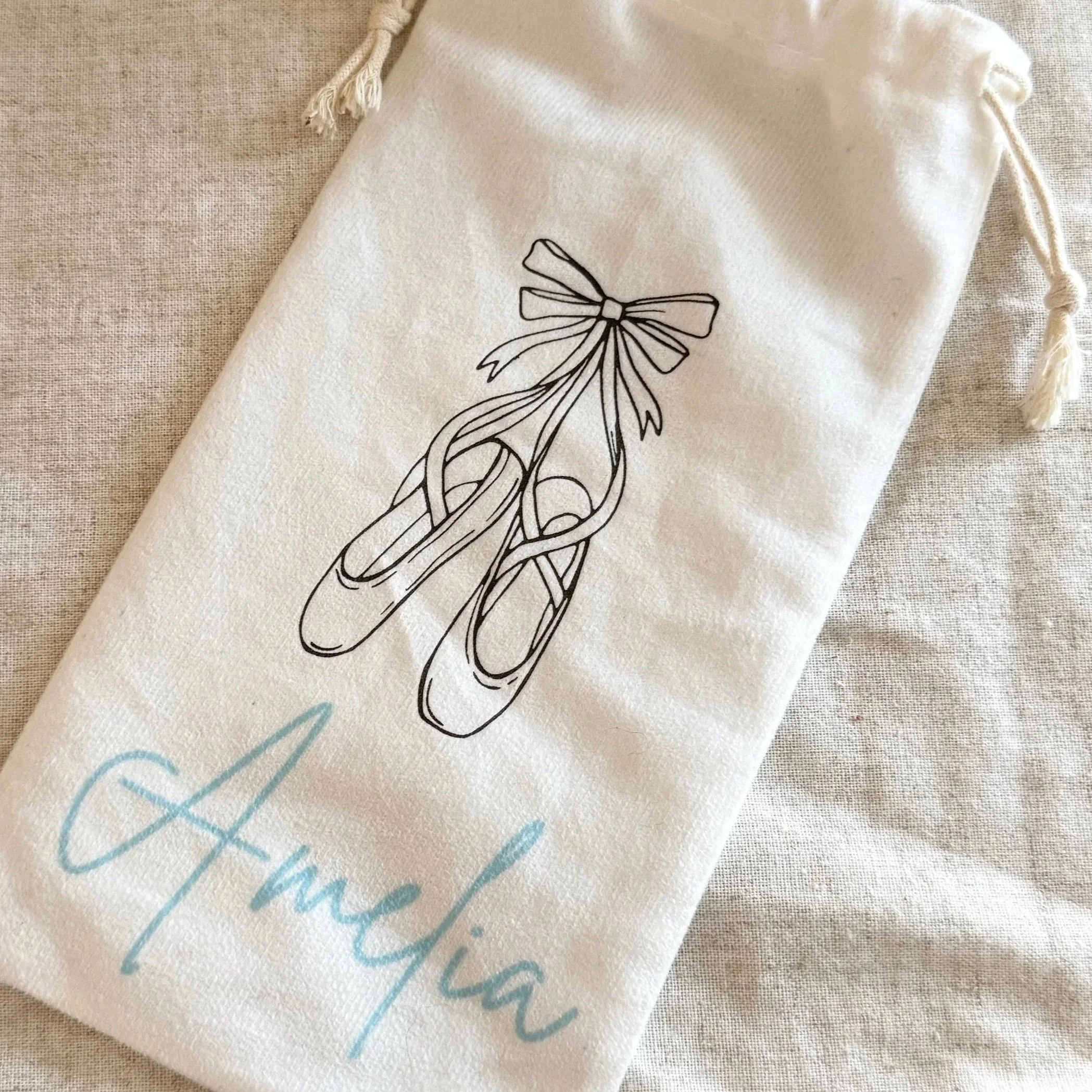 A soft tan colored drawstring bag with a drawing of ballet slippers tied together with a ribbon and the dancers name written in blue cursive below.