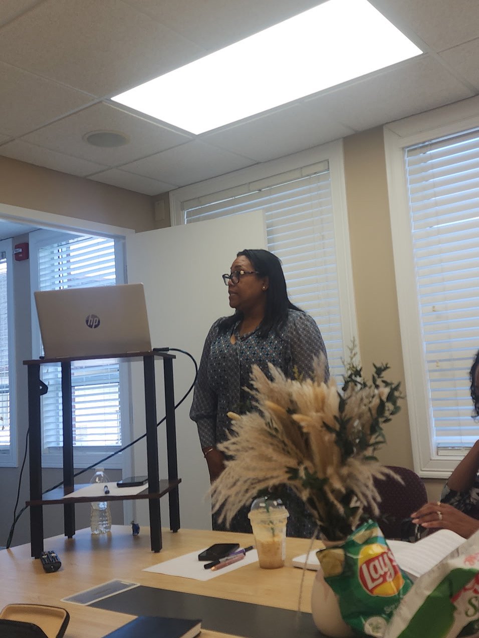 A woman with glasses standing in a meeting room, speaking or presenting, with a laptop on a stand in front of her. The room has windows with blinds, a table with various items including a drink, a bag of Lay's chips, and a floral arrangement.