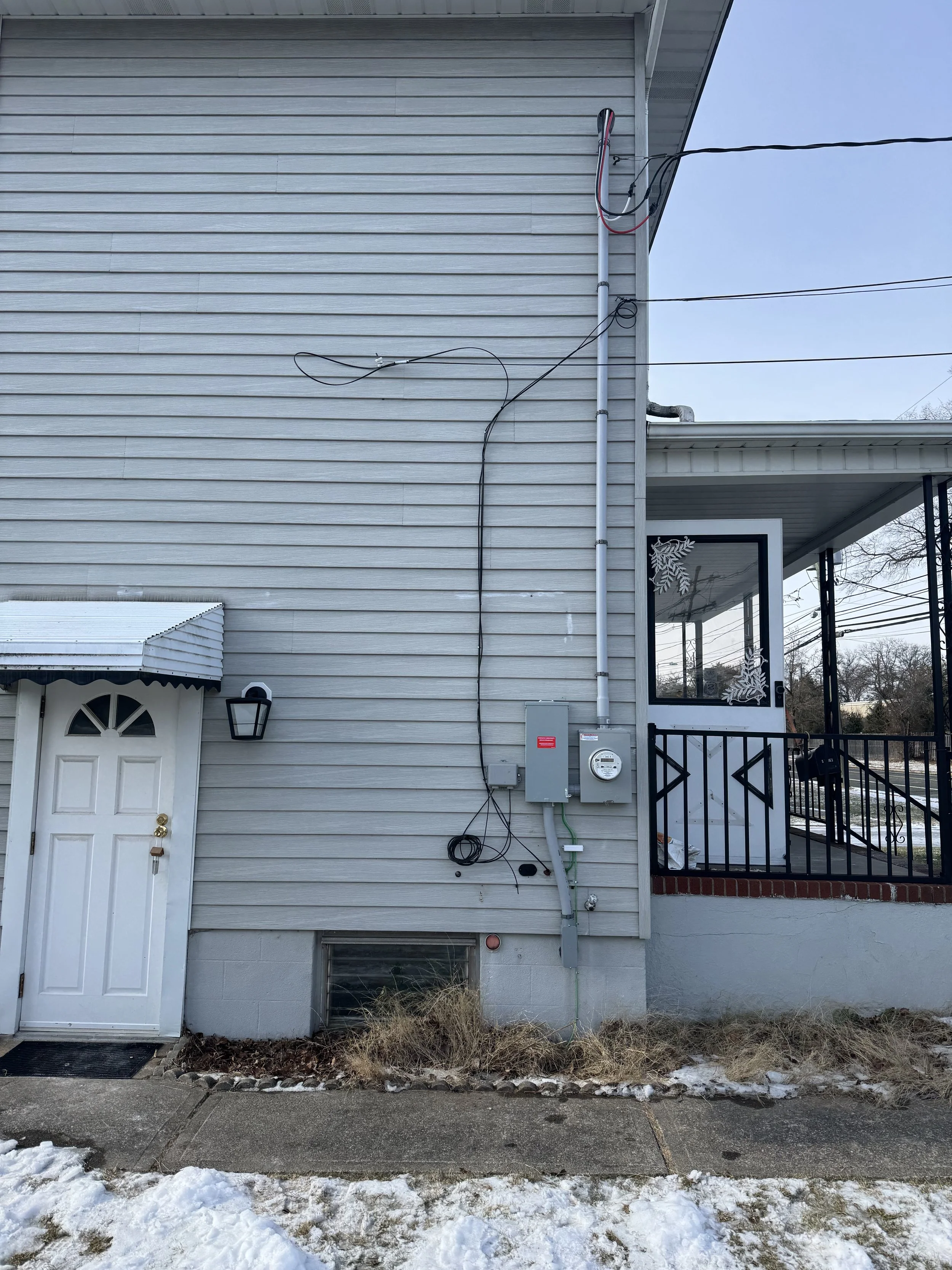 Side view of a house with beige vinyl siding, a small outdoor porch with a black railing, and a white media box near an electrical meter and gray electrical panel. Snow and dead grass on the ground.