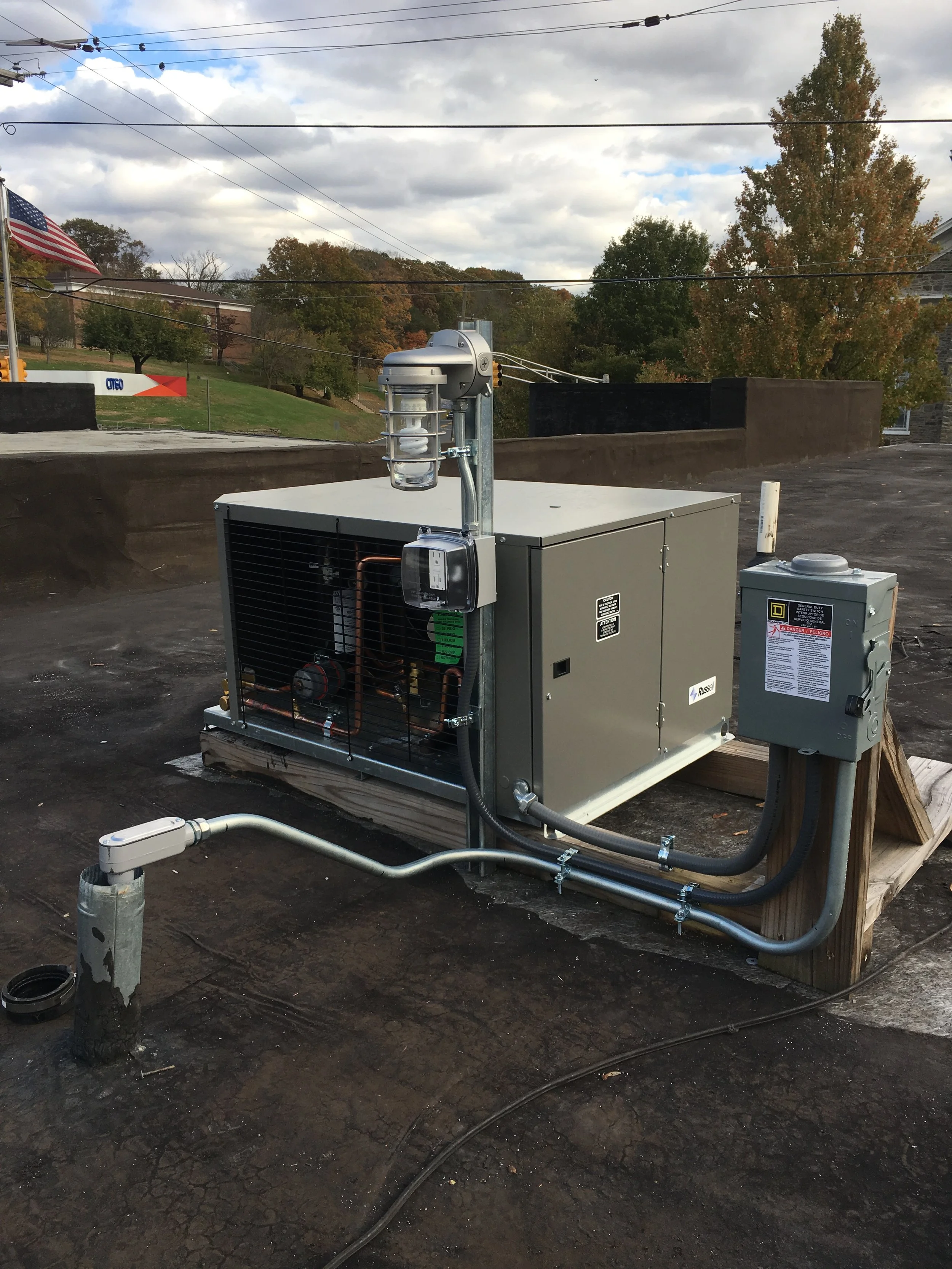 Rooftop HVAC unit on a flat roof with electrical controls, copper piping, and a pressure regulator, with trees and cloudy sky in the background.