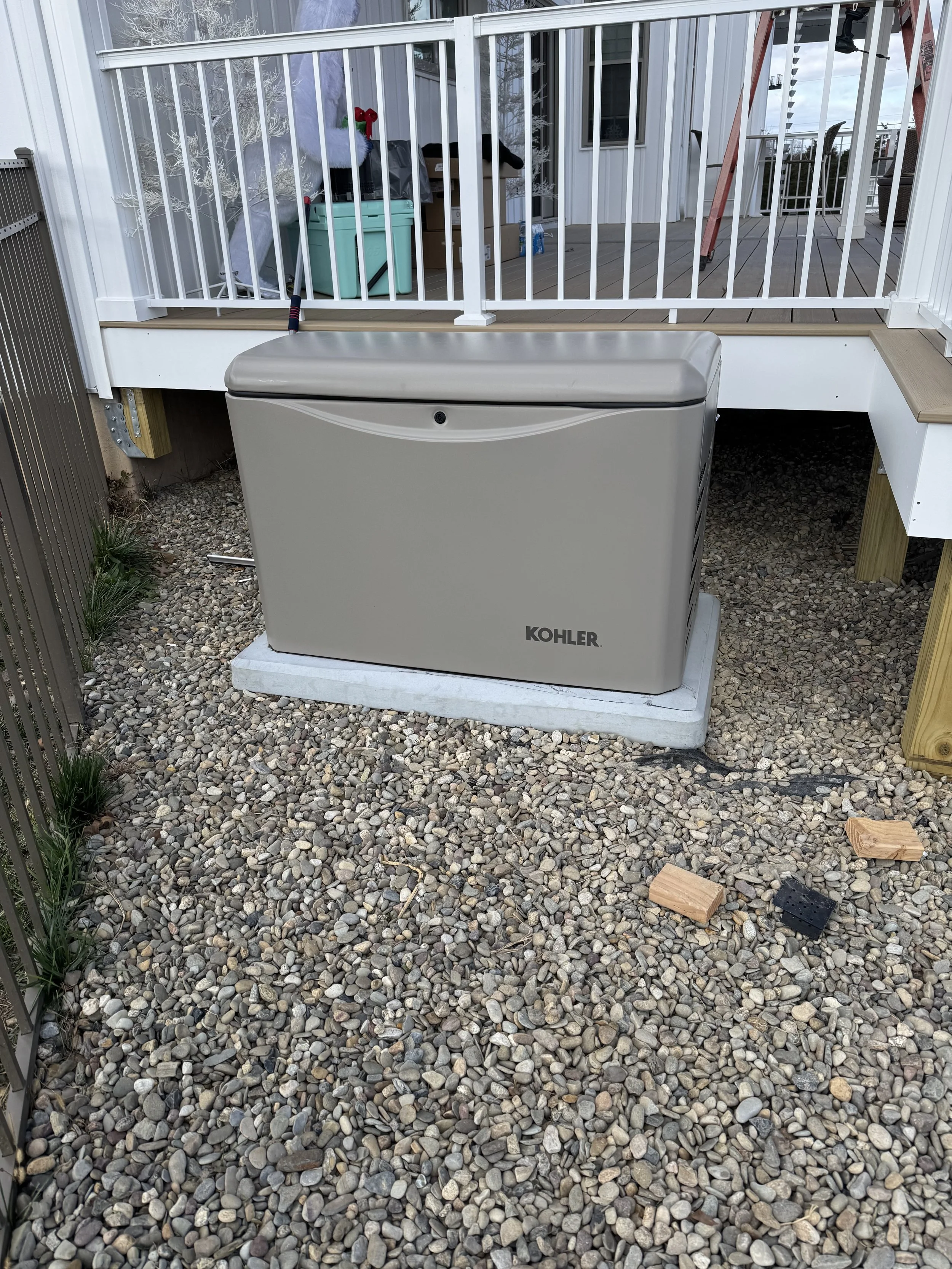 Outside view of a Kohler generator installed on a gravel yard, positioned on a concrete pad beneath a raised deck with white railing, with some wooden blocks and a small black object on the gravel.