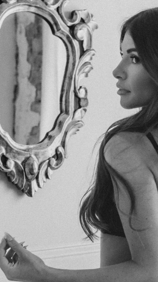 Black and white photo of a woman with long hair looking at a decorative mirror on the wall.