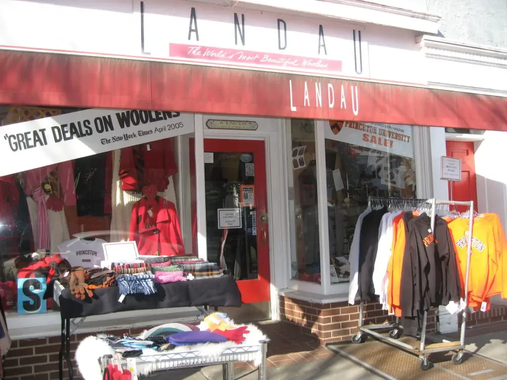 Storefront of Landau clothing retail store with clothing racks outside, including Princeton apparel and scarves, and a window display featuring mannequins in red clothing.