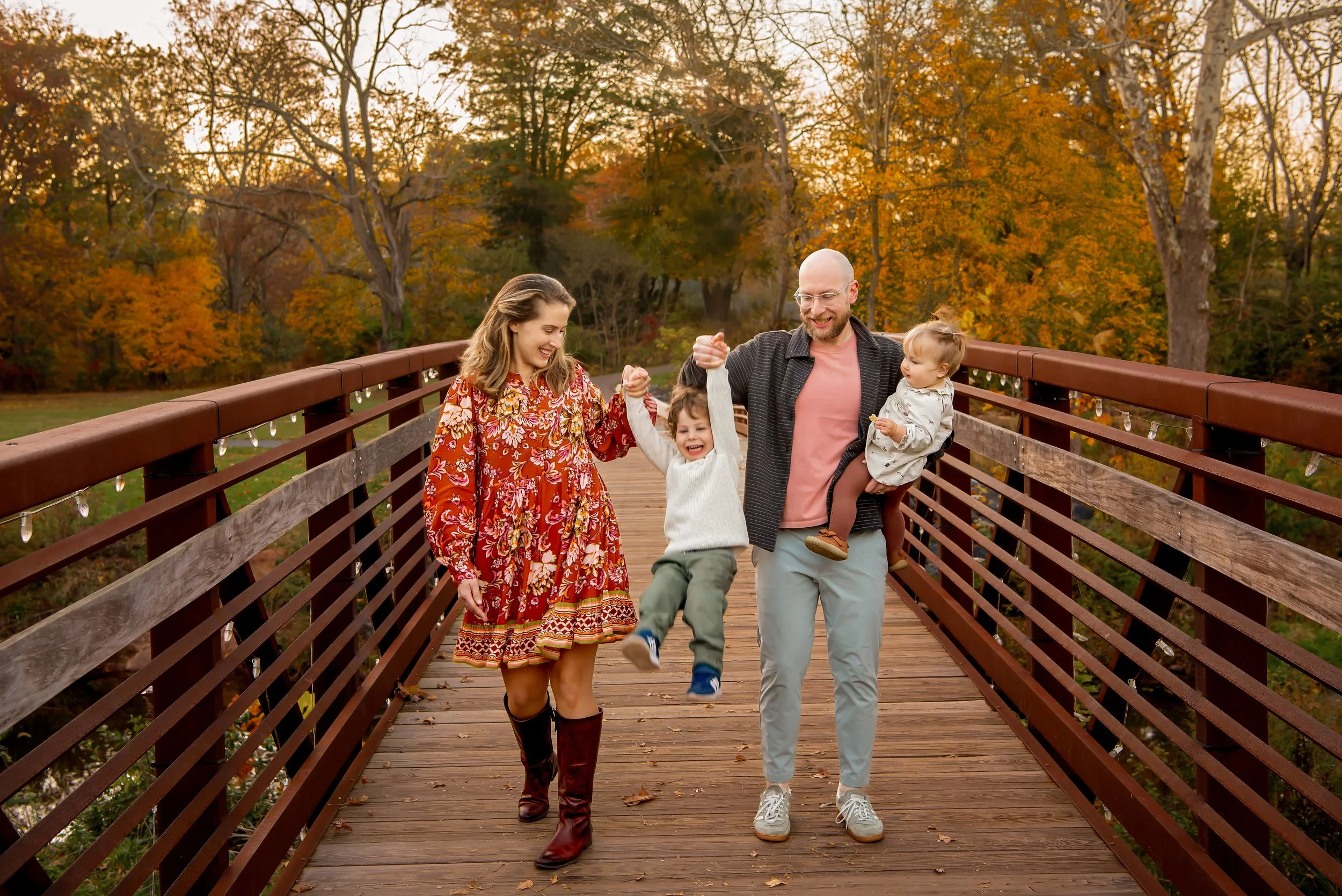 A family of four enjoys walking on a wooden bridge in a park during autumn, with trees with colorful fall leaves in the background.