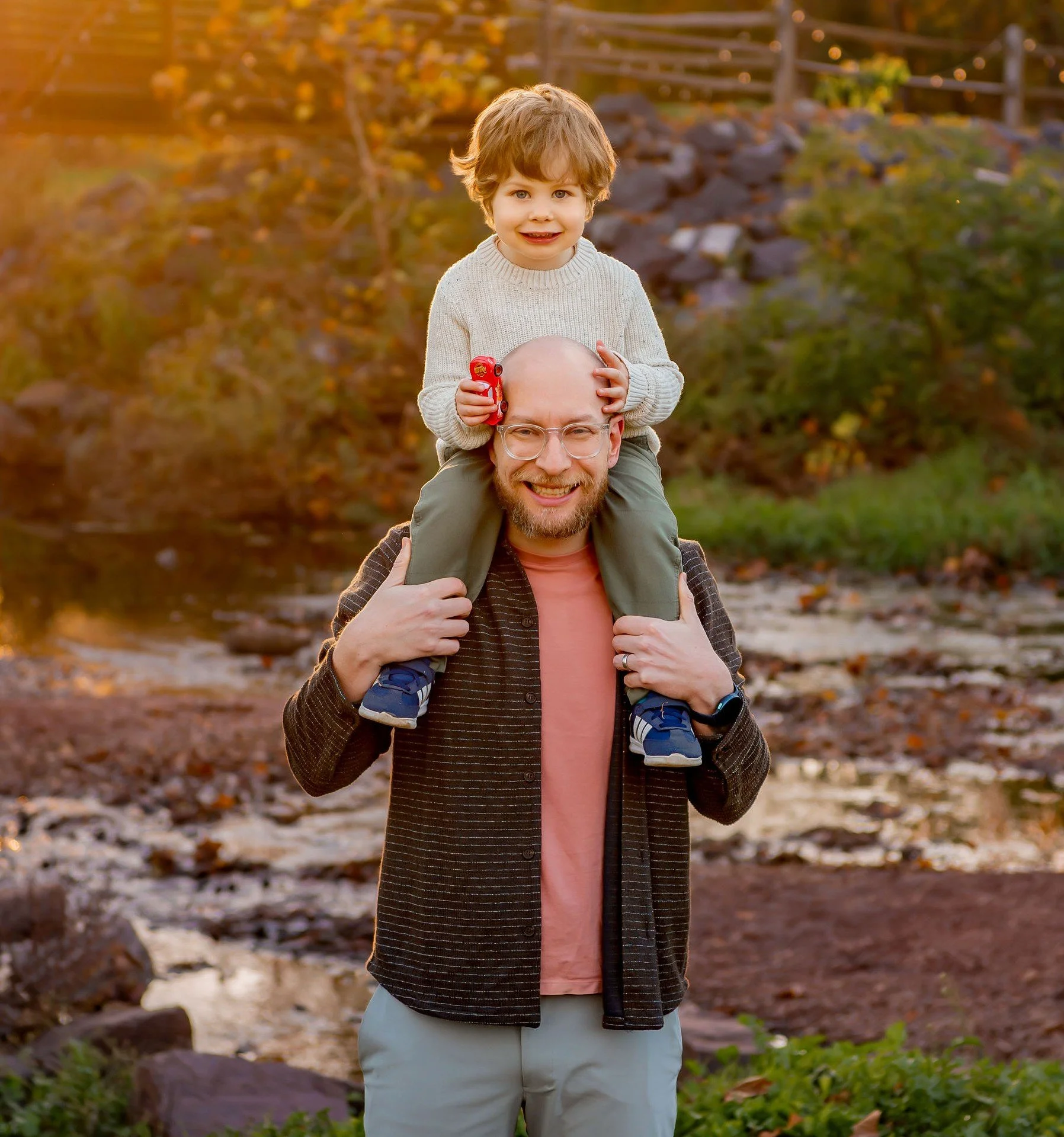 A man with glasses and a beard carrying a young boy on his shoulders in an outdoor park during autumn; the boy is holding a red toy car and smiling.