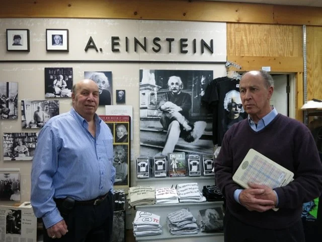 Two men standing in front of a display about Albert Einstein, with photos and memorabilia, one holding a booklet.