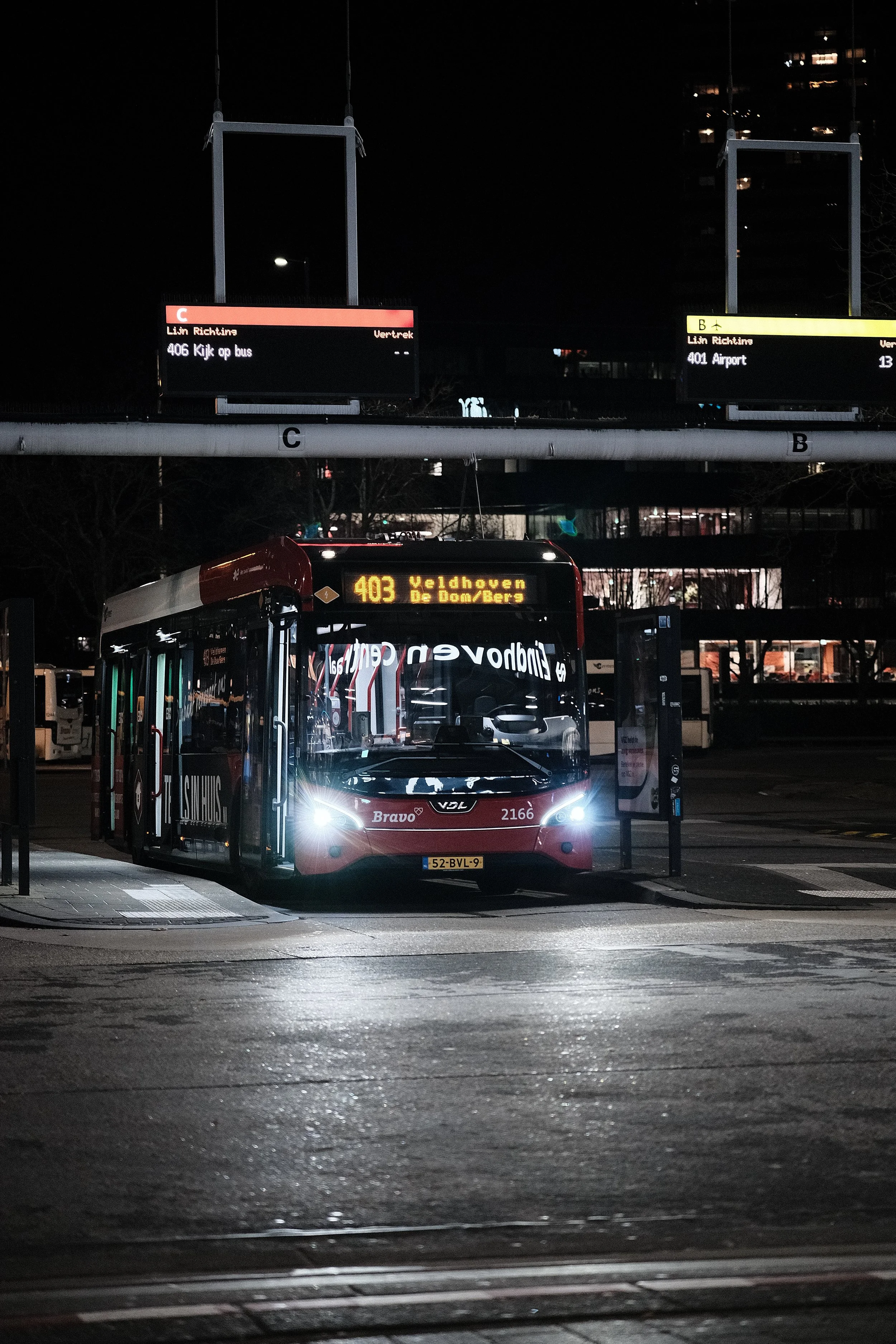 A city bus stopped at a bus stop at night, with its lights on and digital display showing route 403 to Veldhoven De Dom/Bergs.