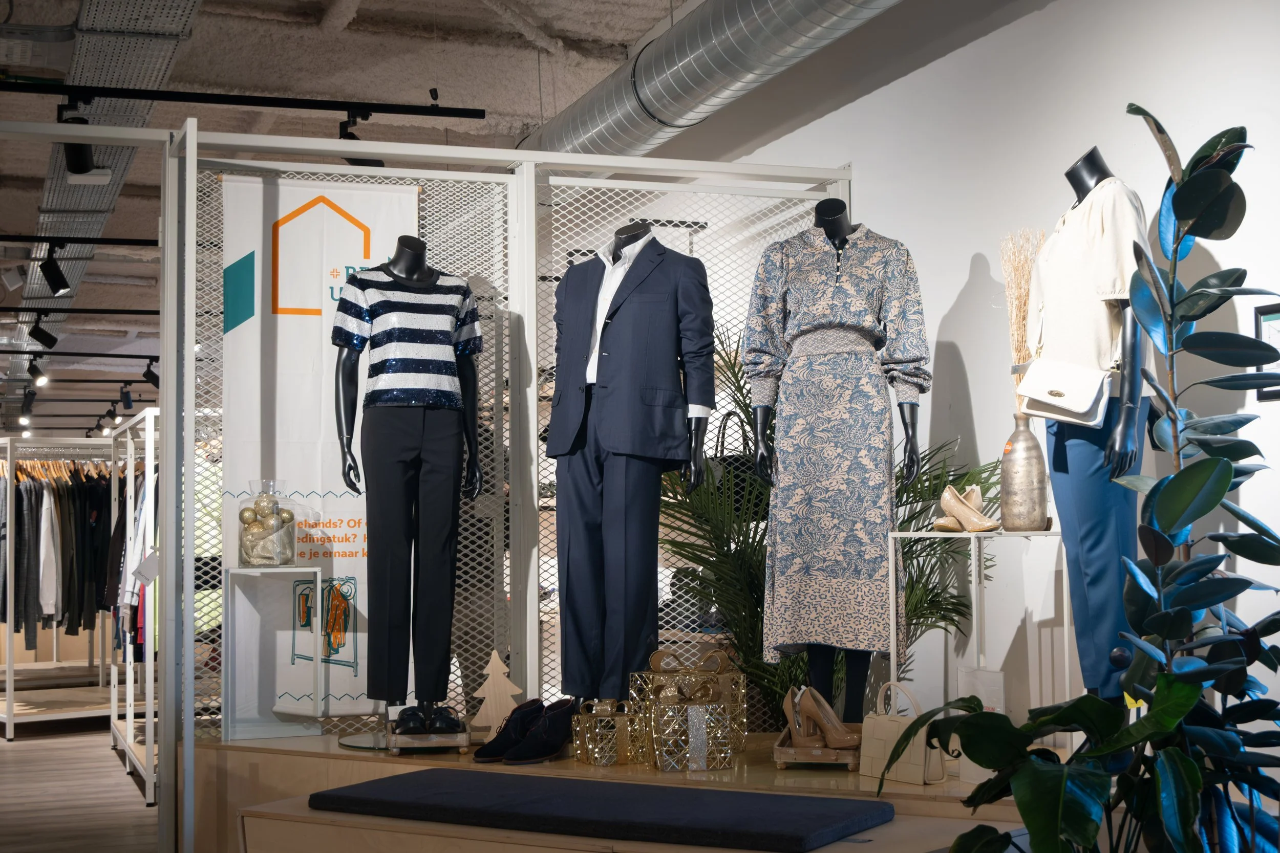 Display of mannequins dressed in fashionable clothing, including striped top, navy suit, patterned dress, and casual outfit, surrounded by decorative plants and holiday ornaments at a retail store