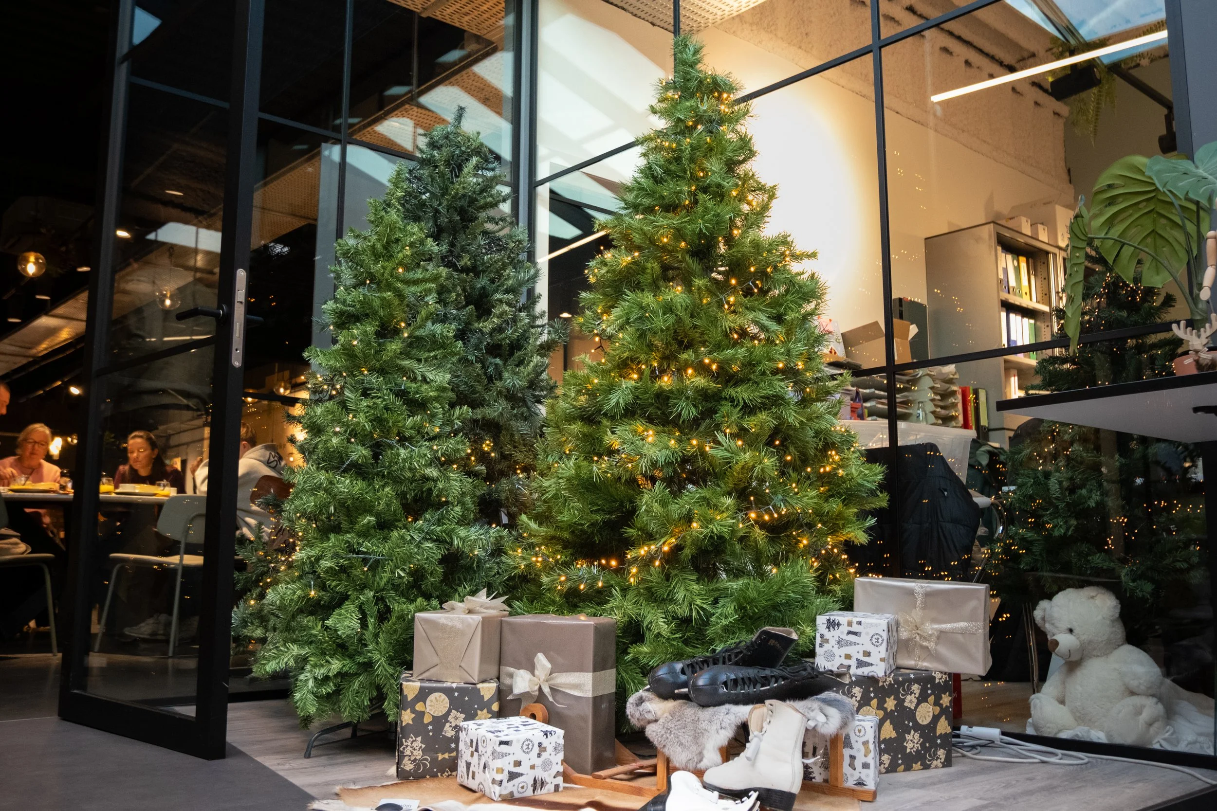 Indoor holiday scene with two decorated Christmas trees with lights, wrapped gifts, a teddy bear, and winter shoes, viewed through glass walls with people dining inside.