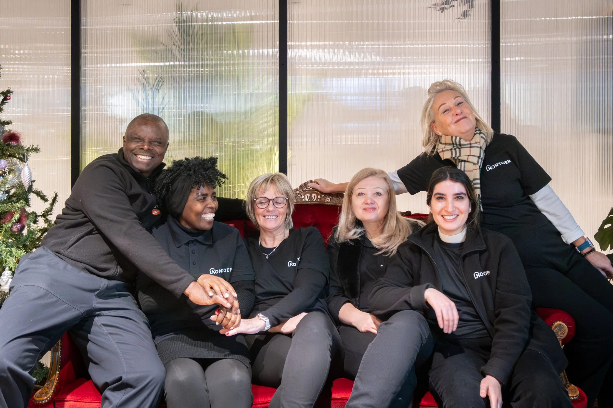 A group of six diverse smiling people, five women and one man, sitting and standing around a red velvet sofa with a Christmas tree visible on the side, in a well-lit indoor setting.