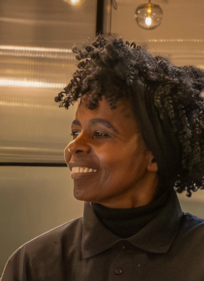 A woman with dark curly hair smiling indoors, wearing a black collared shirt.