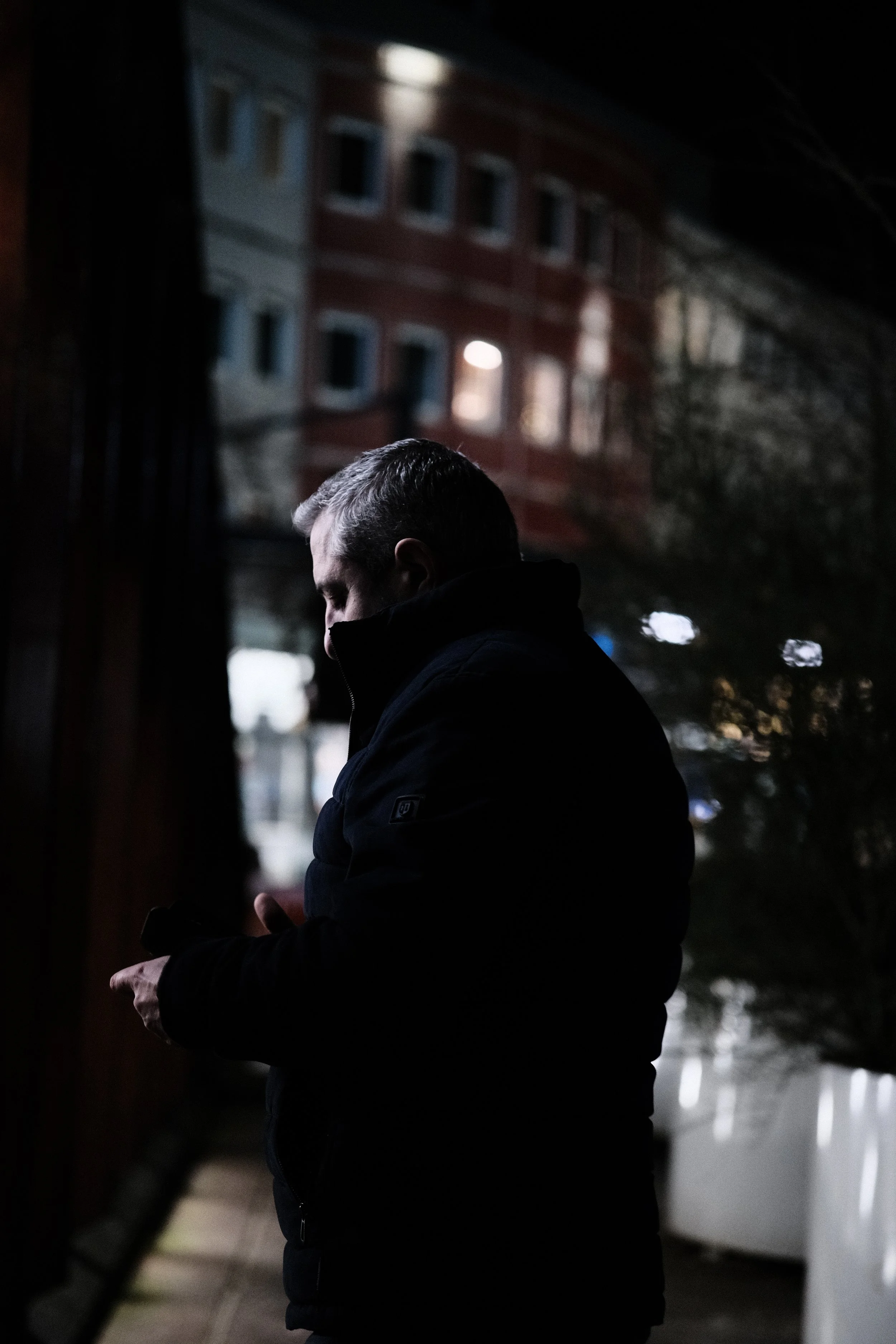 A man in a dark jacket standing outside at night, looking and using his smartphone.