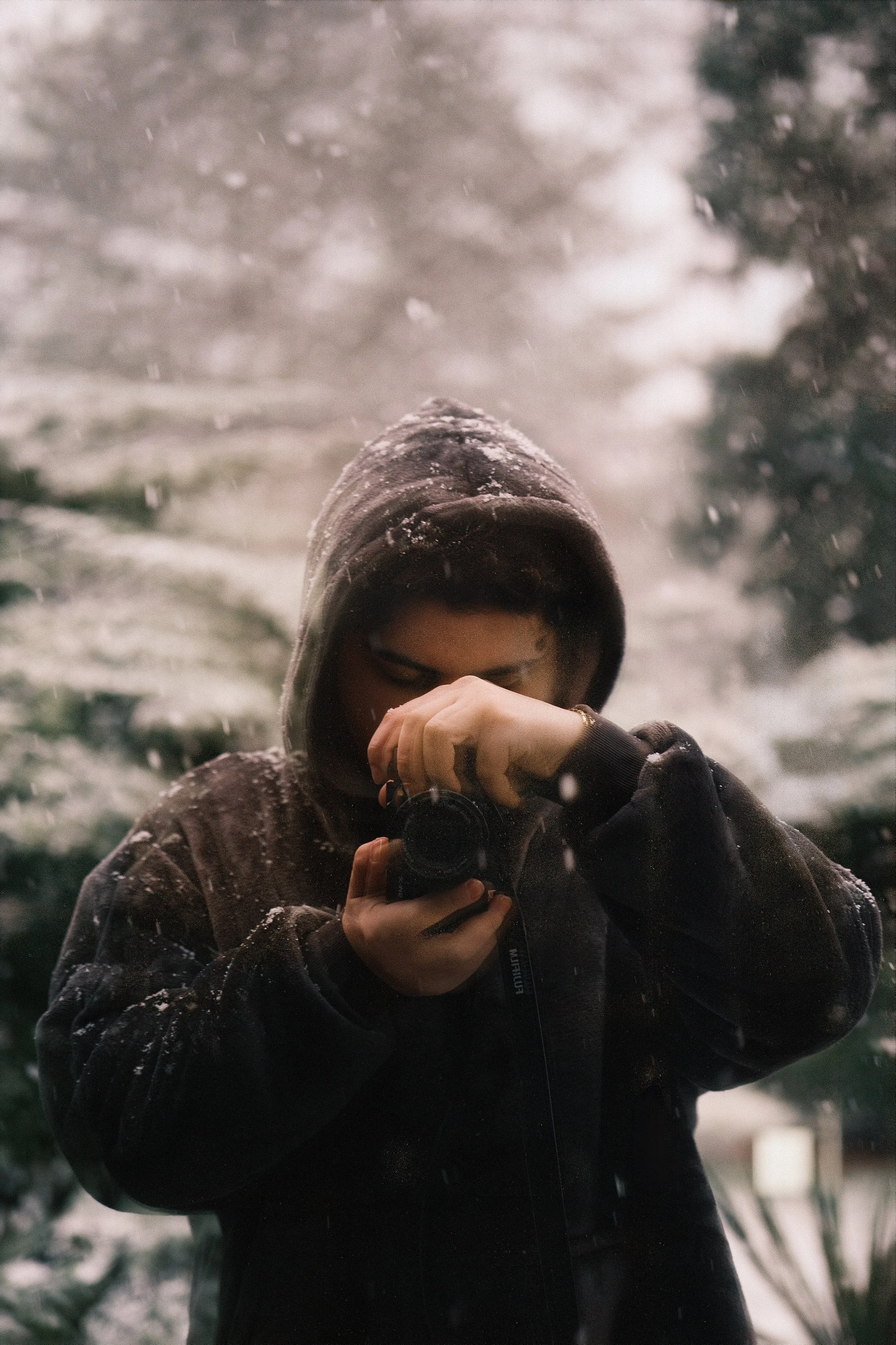 Person taking a photo with a camera outside during snowfall, wearing a hooded jacket.