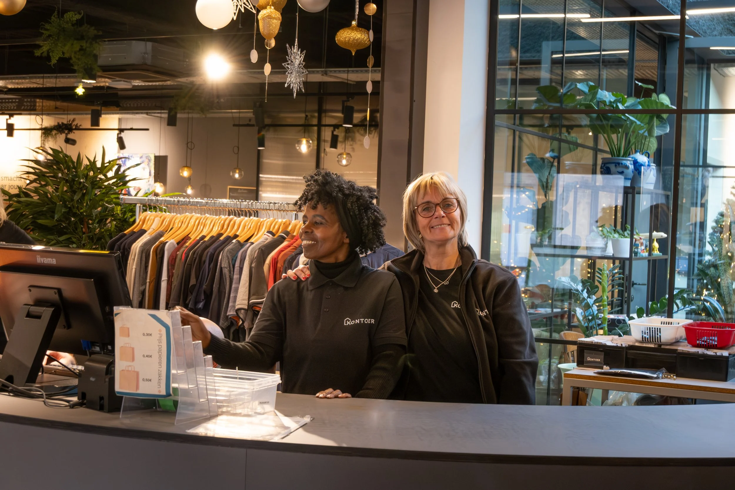 Two women working at a retail checkout counter surrounded by clothing racks and decorative hanging ornaments, with large window and green plants in the background.