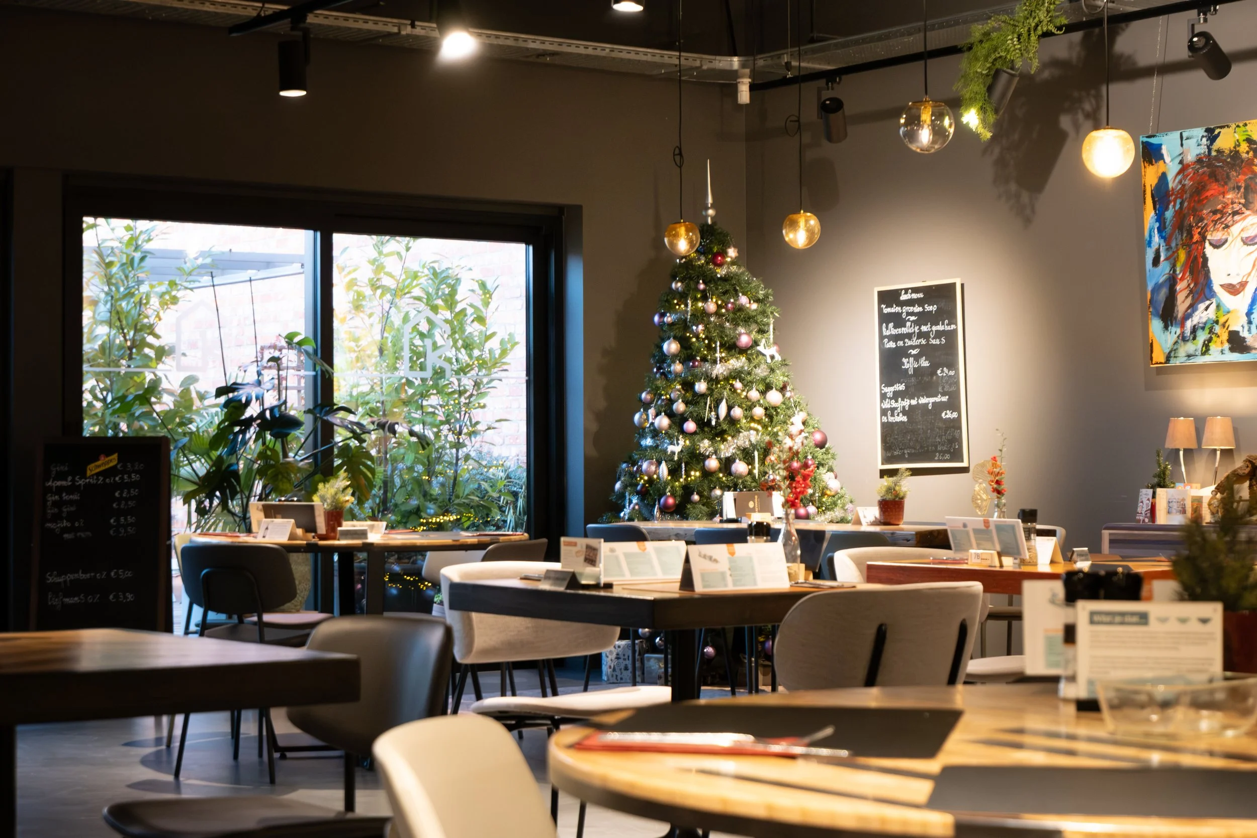 Interior of a cafe decorated for Christmas with a Christmas tree, hanging lights, tables, chairs, and large window with plants outside.