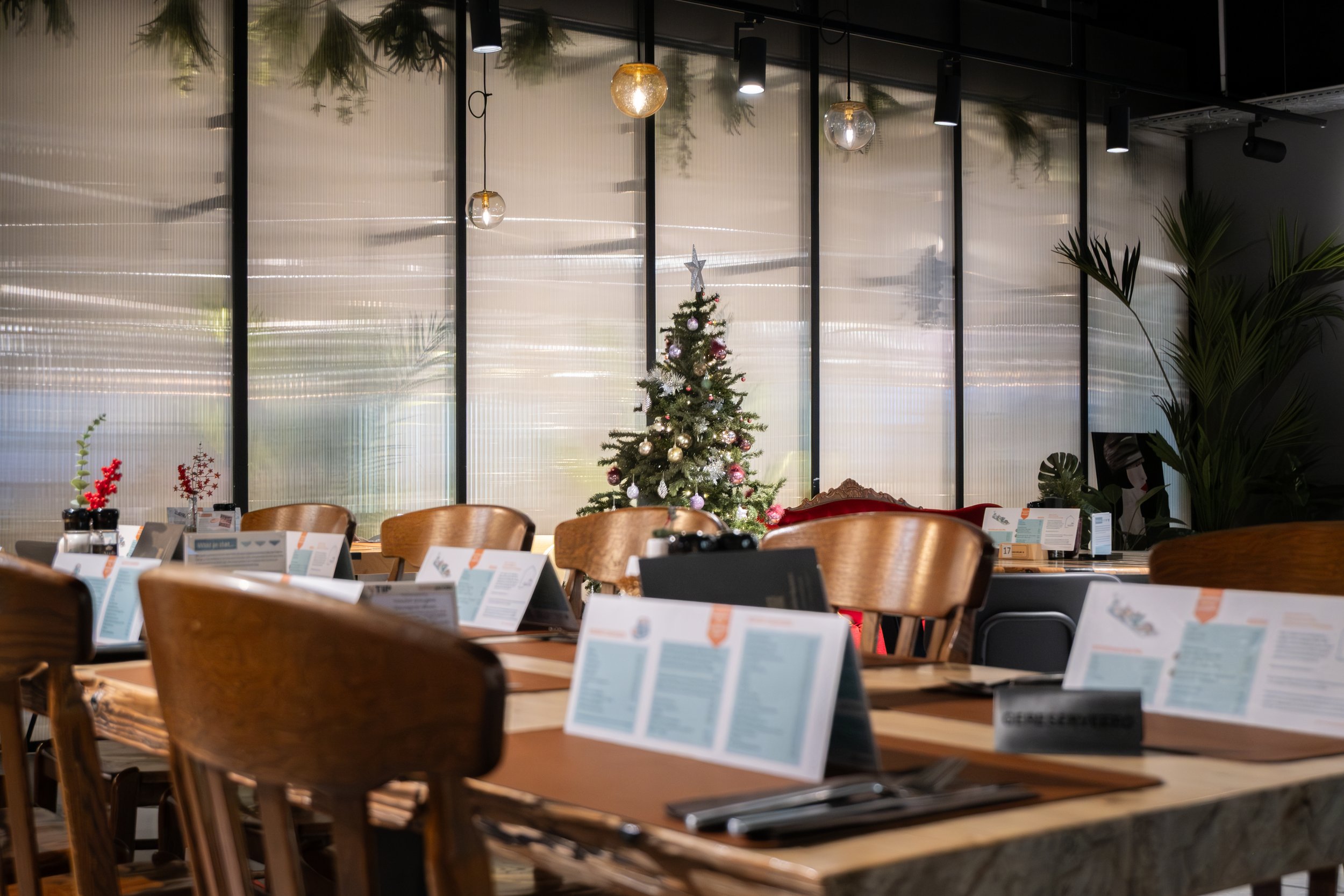 A decorated Christmas tree in a modern restaurant with wooden chairs and tables, menu cards, and green plants in the background.