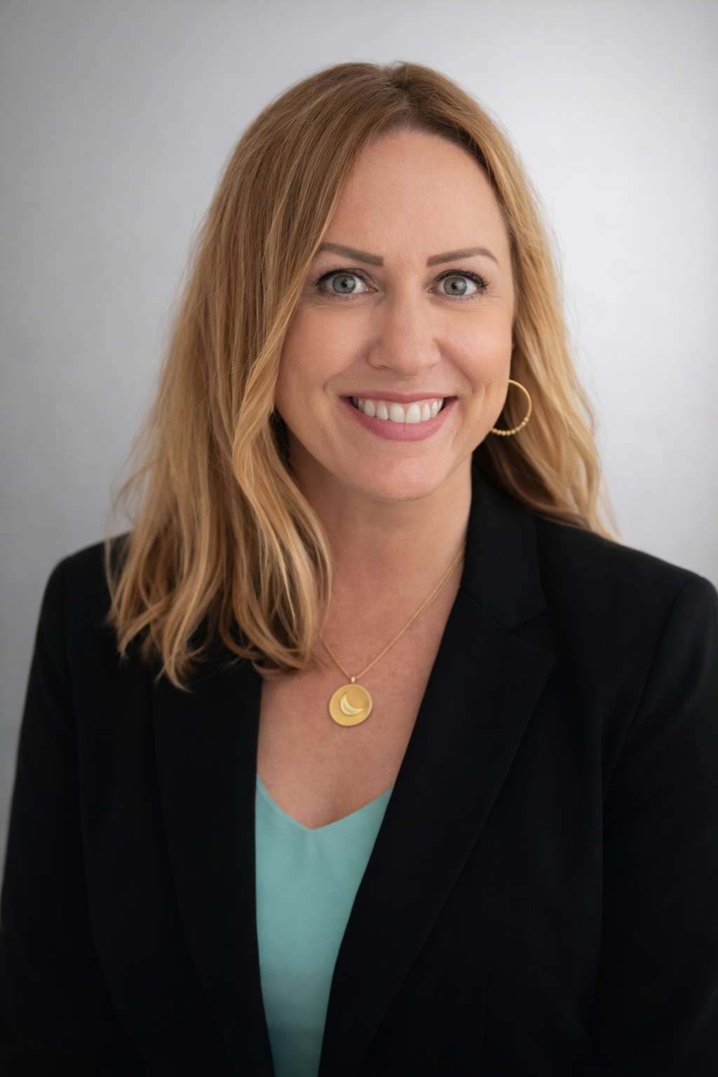 A woman with blonde hair wearing a black blazer, gold hoop earring, and gold moon necklace, smiling at the camera against a plain background.