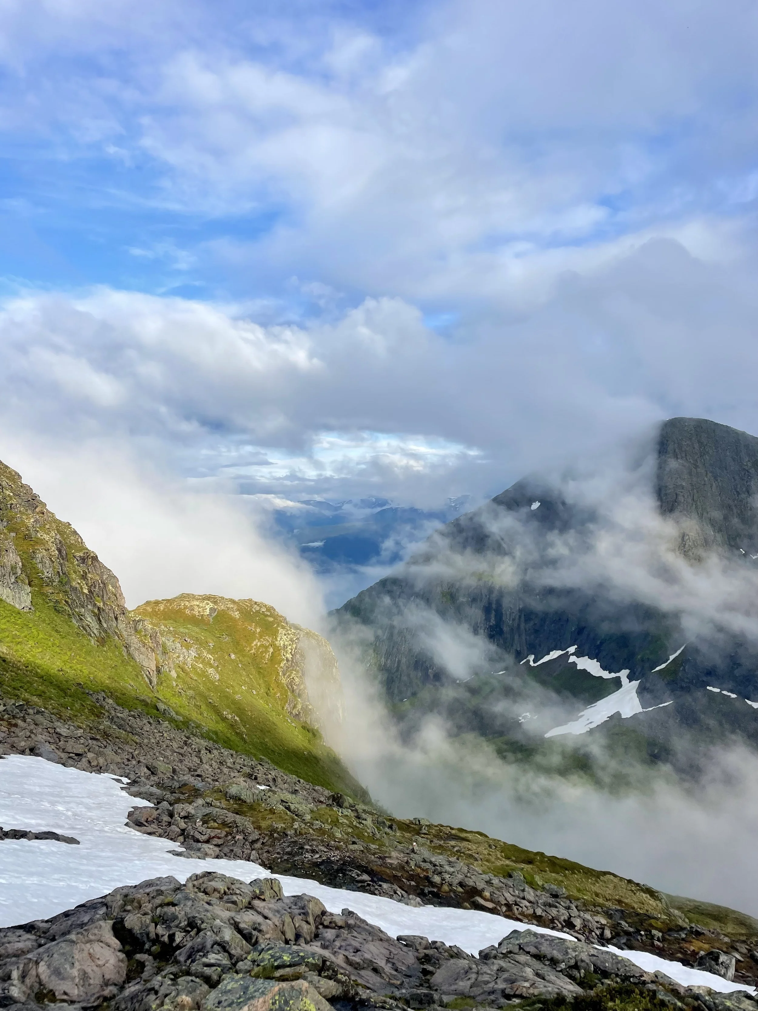 Scenic mountain landscape with rocky, mossy slopes, patches of snow, and clouds in the sky, with distant mountains and fog.