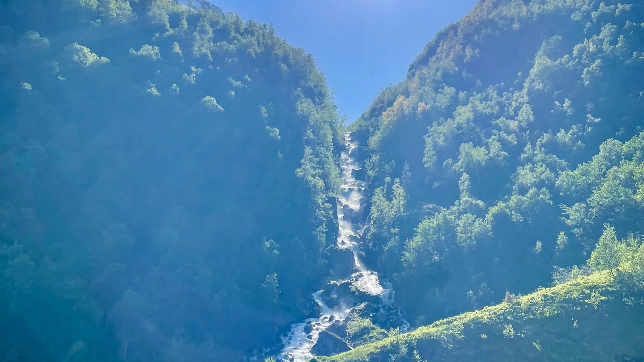 Waterfall cascading down a mountain surrounded by dense green trees on a clear, sunny day.