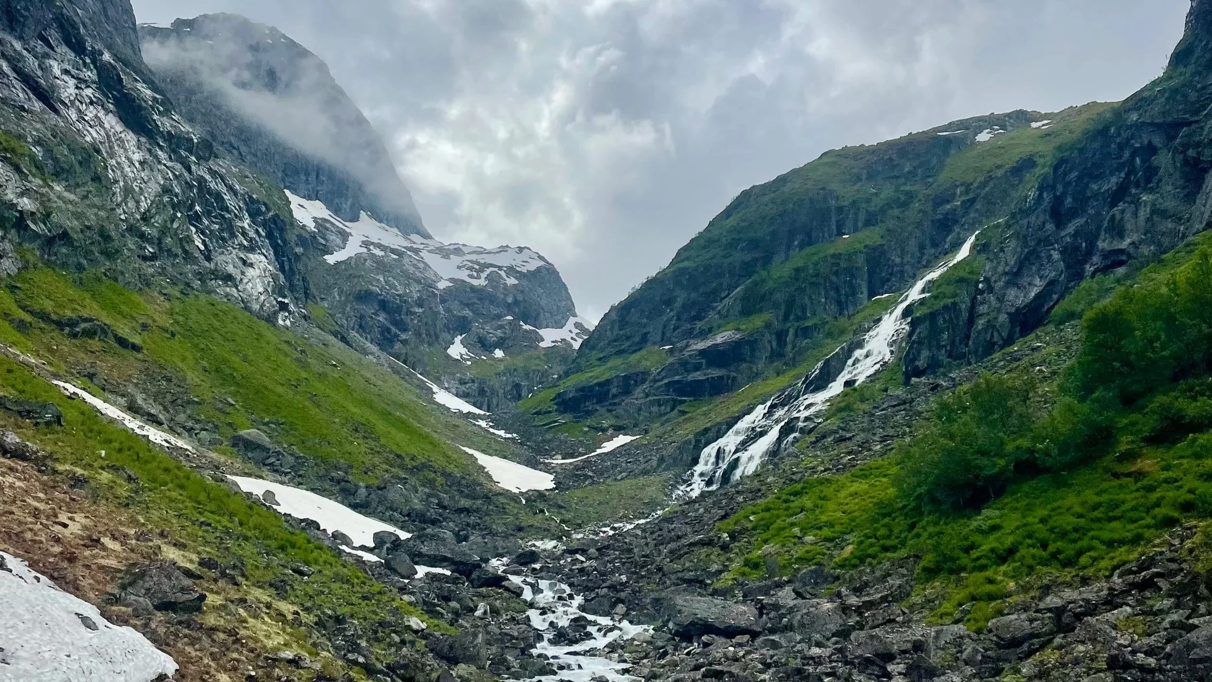 Mountain valley with snow patches, green slopes, rocky terrain, and waterfalls under cloudy sky.