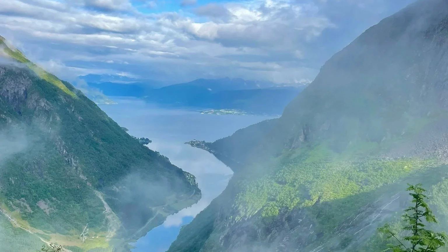 A scenic view of a fjord with steep, green mountains on either side, and a calm river flowing through the valley under a cloudy sky.