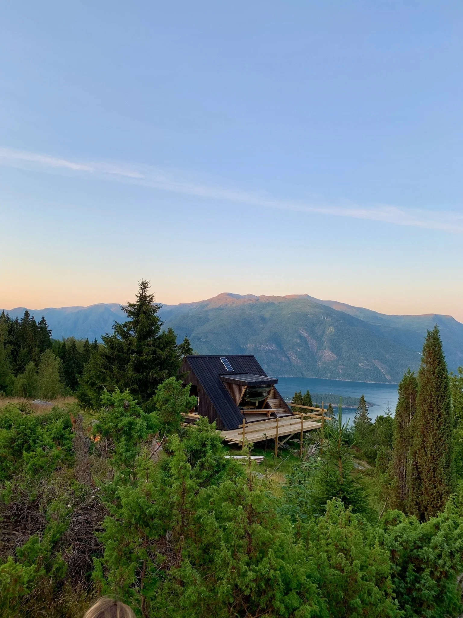 A small wooden house with solar panels on the roof, situated in a lush green forested hillside, overlooking a large body of water and mountains during sunset.