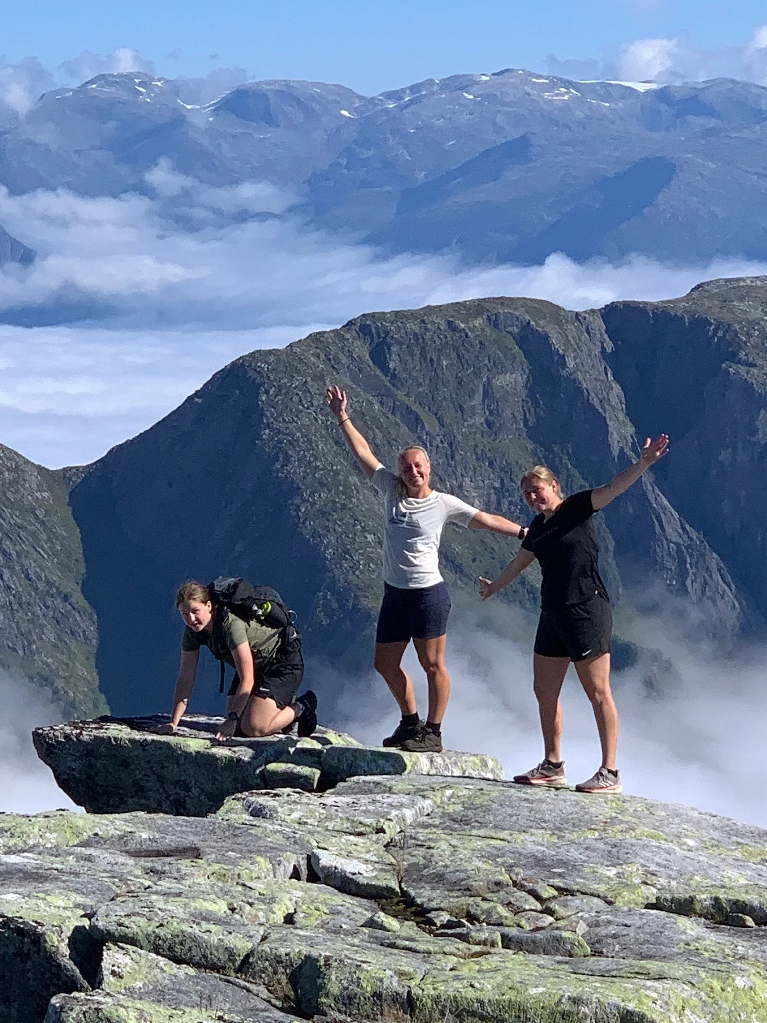 Three hikers on a rocky mountain peak with mountain ranges and clouds in the background.