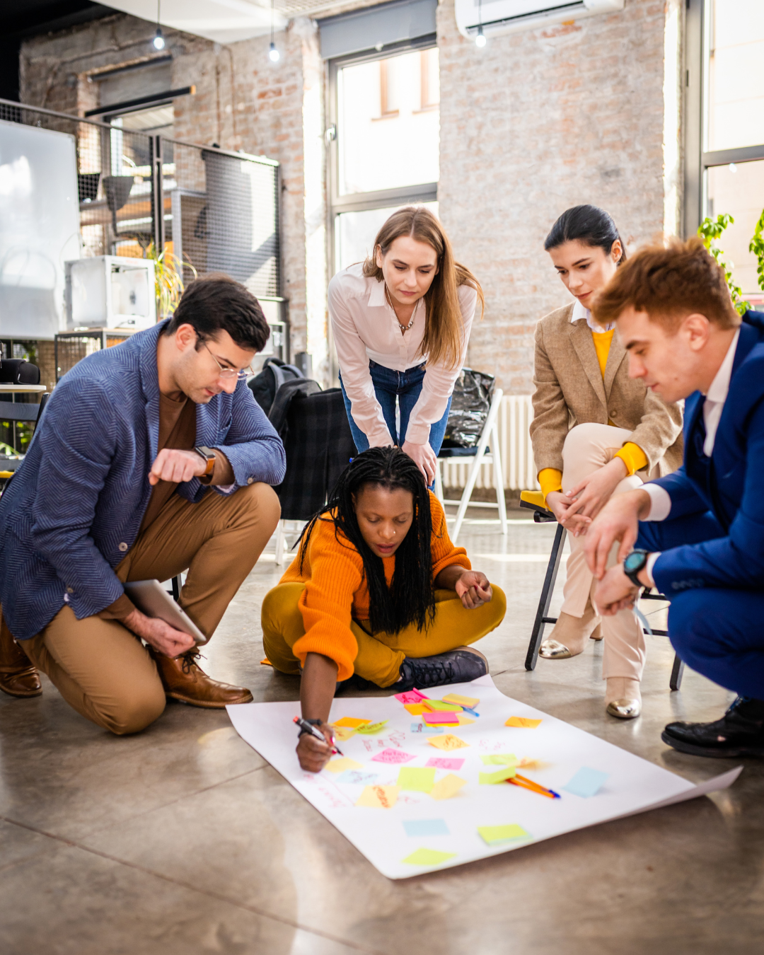 A diverse group of five young adults collaborating on a project in a modern, industrial-style office with large windows. Some are kneeling or sitting on the floor around a large sheet of paper with colorful sticky notes and markers, actively discussing and writing ideas.
