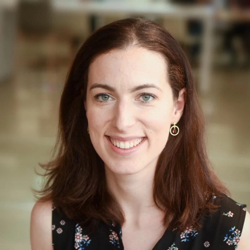 Portrait of a woman with shoulder-length brown hair, green eyes, and earrings, smiling indoors with a blurred background.
