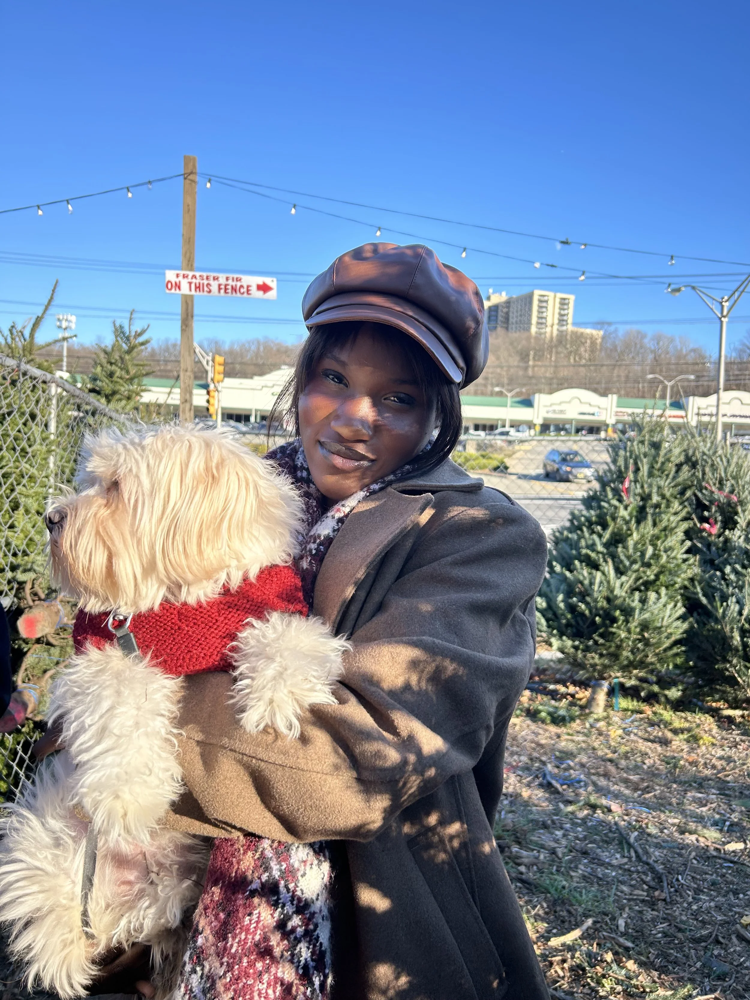 A woman in a black leather hat and dark coat holding a small, fluffy dog wearing a red sweater outdoors on a sunny day. There are trees, a chain-link fence, and a parking lot in the background.