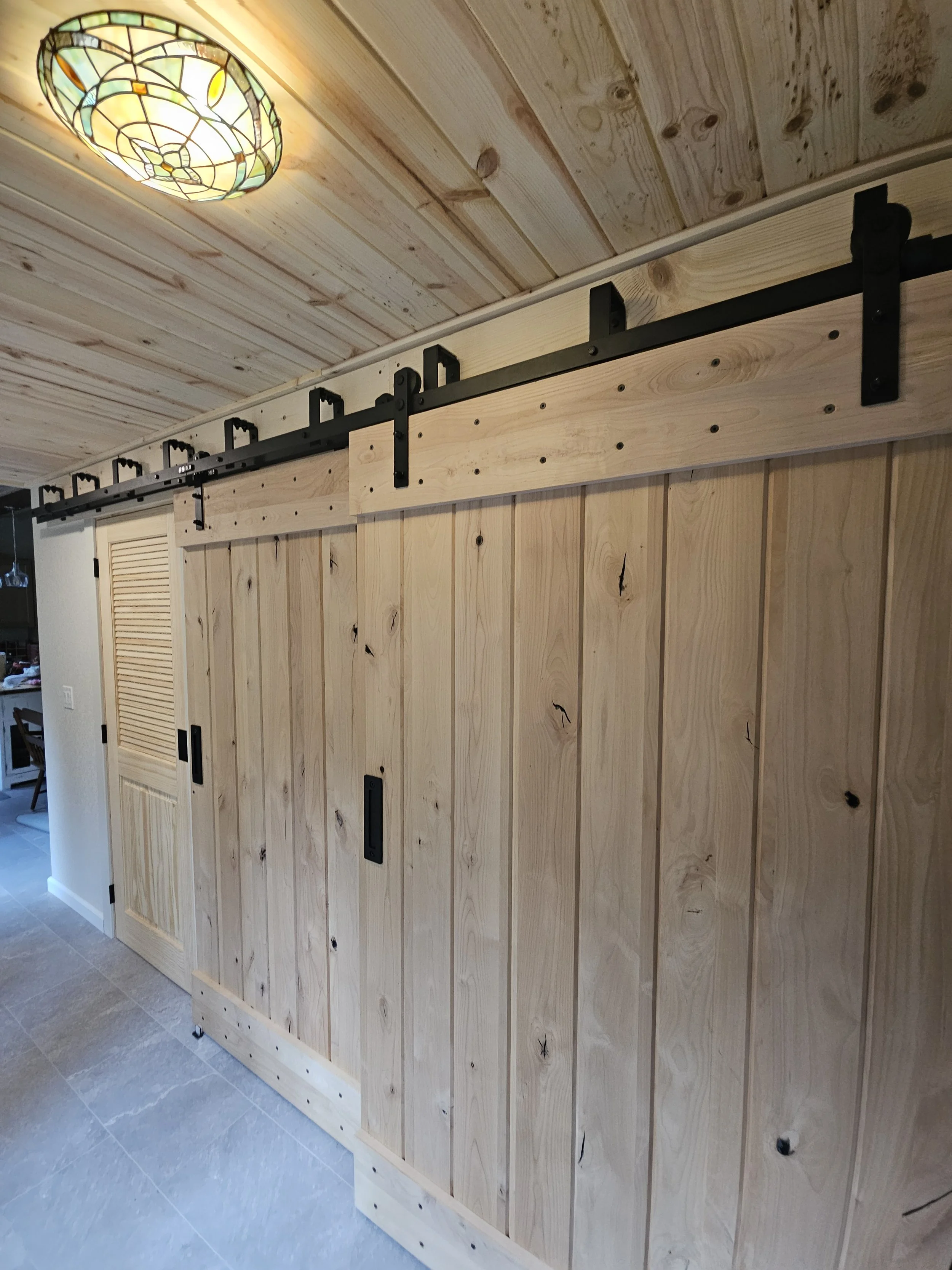 A wooden sliding barn door on a black metal track system inside a house, with a stained glass ceiling light in a room with wood paneled walls and tile flooring.